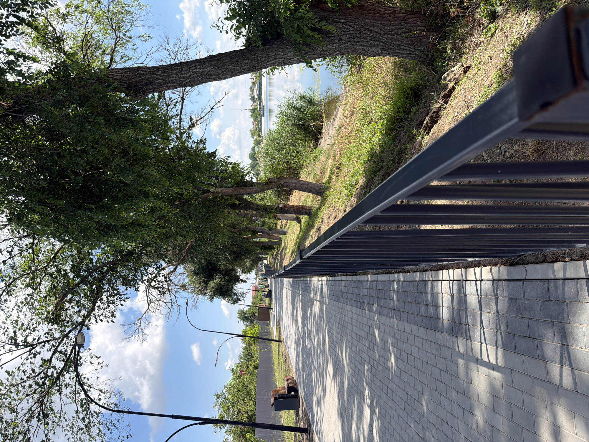 This image depicts a paved walkway in Leningradskaya, Russia, running alongside a body of water bordered by trees and vegetation. The walkway is constructed of light-colored rectangular pavers, with shadows cast across it from overhead trees. A dark metal railing runs along the edge of the walkway, separating it from a grassy embankment leading down to the water. Several streetlights are visible along the walkway, and a bench and trash receptacle are positioned on the left side of the path. The water is calm, reflecting the blue sky and scattered white clouds. On the far side of the water, buildings and more greenery are visible. The overall impression is that of a well-maintained public area, likely a park or promenade.