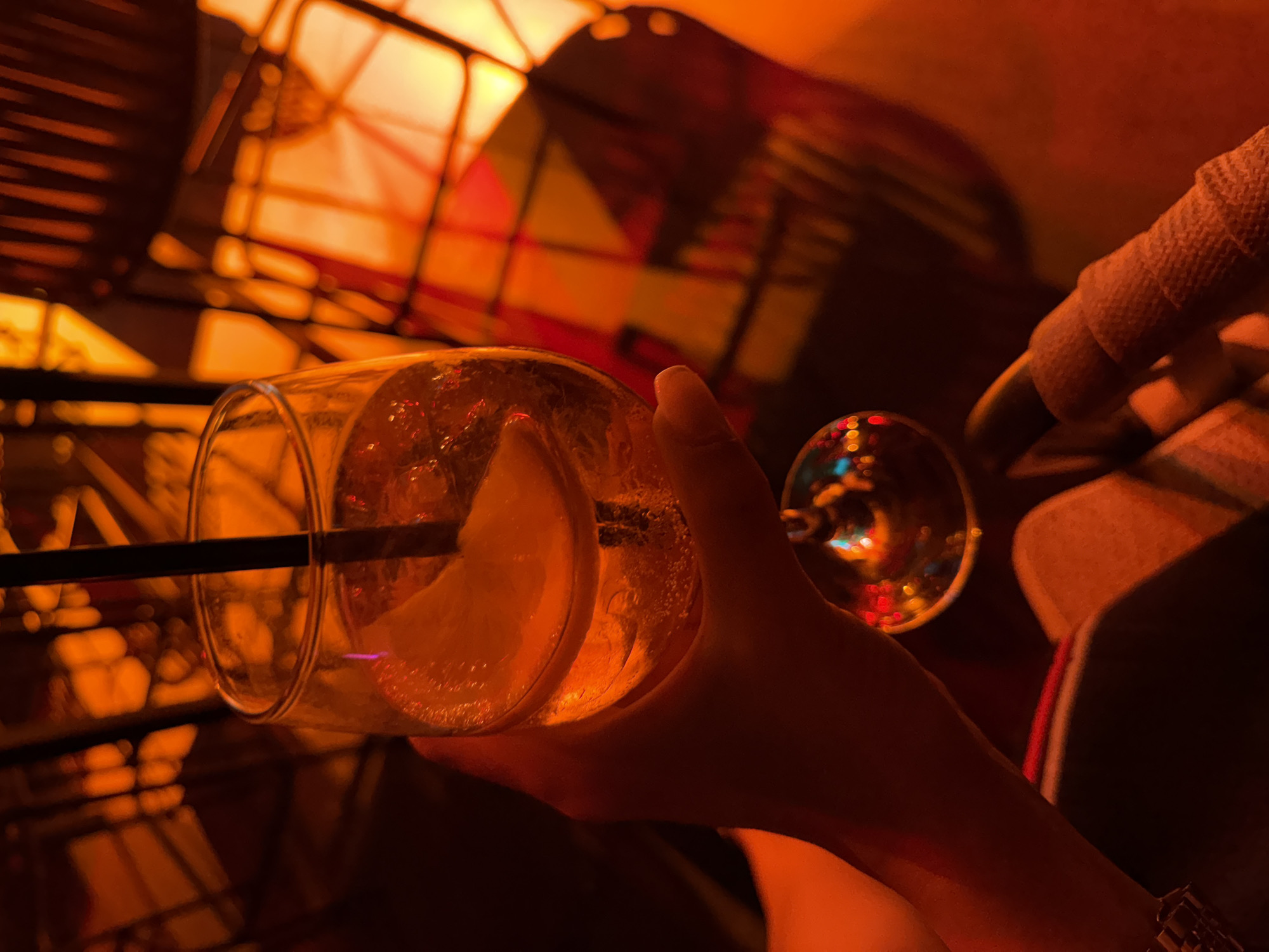 A hand holds a stemmed glass filled with an amber-colored beverage and ice, garnished with a lemon slice and a black straw. The glass is captured in sharp focus, contrasting with the blurred background of a dimly lit establishment in Bodrum, Turkey, illuminated by warm, orange-hued lighting. A decorative, geometric lampshade casts a patterned shadow. In the foreground, another hand with a watch and a bandaged finger is visible. The overall ambiance suggests a relaxed evening at a bar or restaurant.