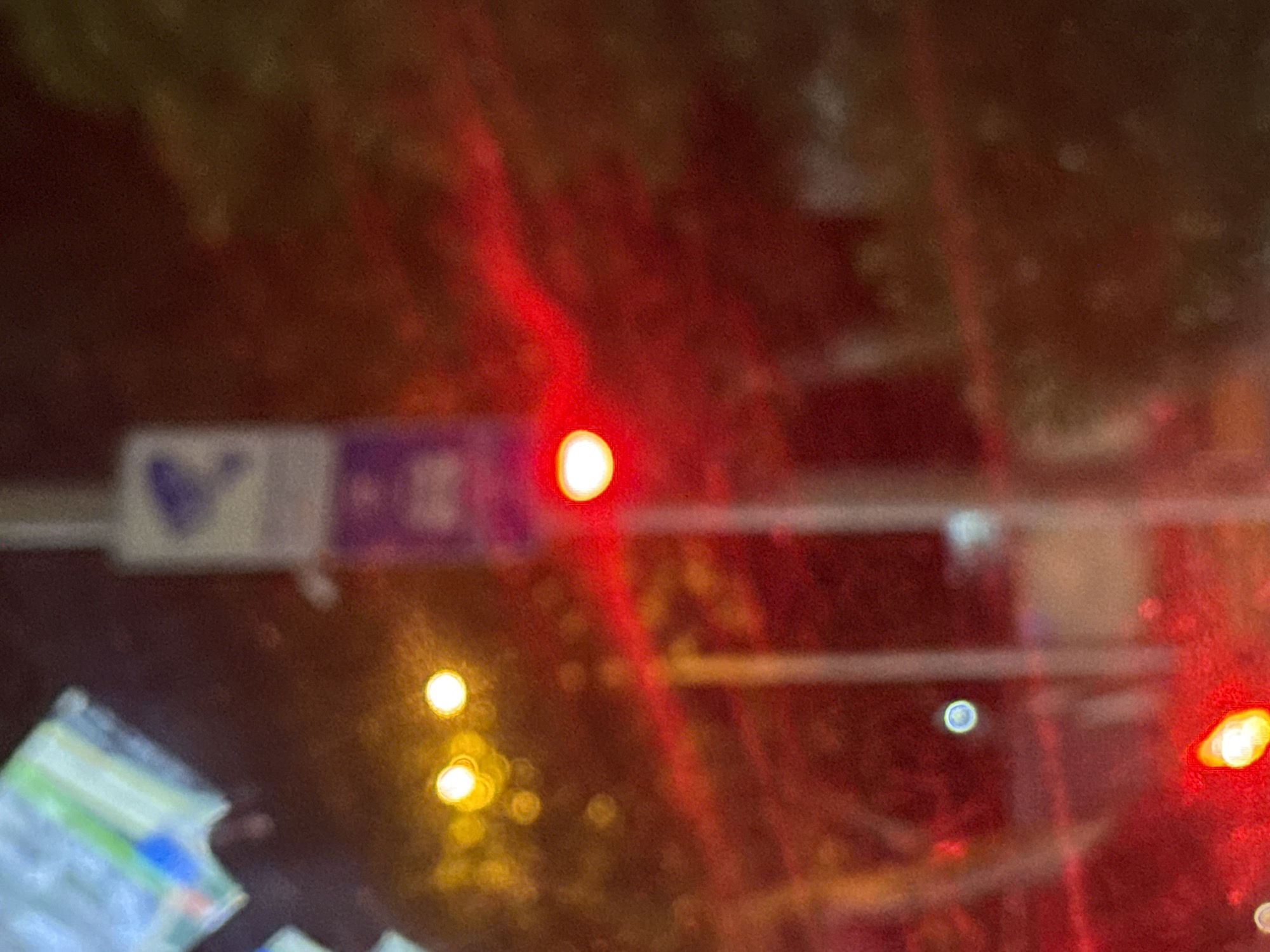 The image shows a blurry, red-tinted view of a street scene at night, likely from inside a vehicle looking out through a wet windshield. In the foreground, a bright red light emanates from what appears to be a traffic signal. Streaks of red light are visible, suggesting motion or reflections on the glass. In the background, two blurry yellow lights are present, possibly streetlights or other vehicle lights. A vertically oriented sign with some blue and purple elements is partially visible on a pole. The overall environment appears to be urban, with trees or foliage visible in the background, also obscured by darkness and blur.
While the image's content does not definitively place it in Tbilisi, Georgia, it is common for traffic lights, street signs, and urban nighttime streetscapes to appear similar across various cities. Without specific identifying markers, a precise location cannot be determined.