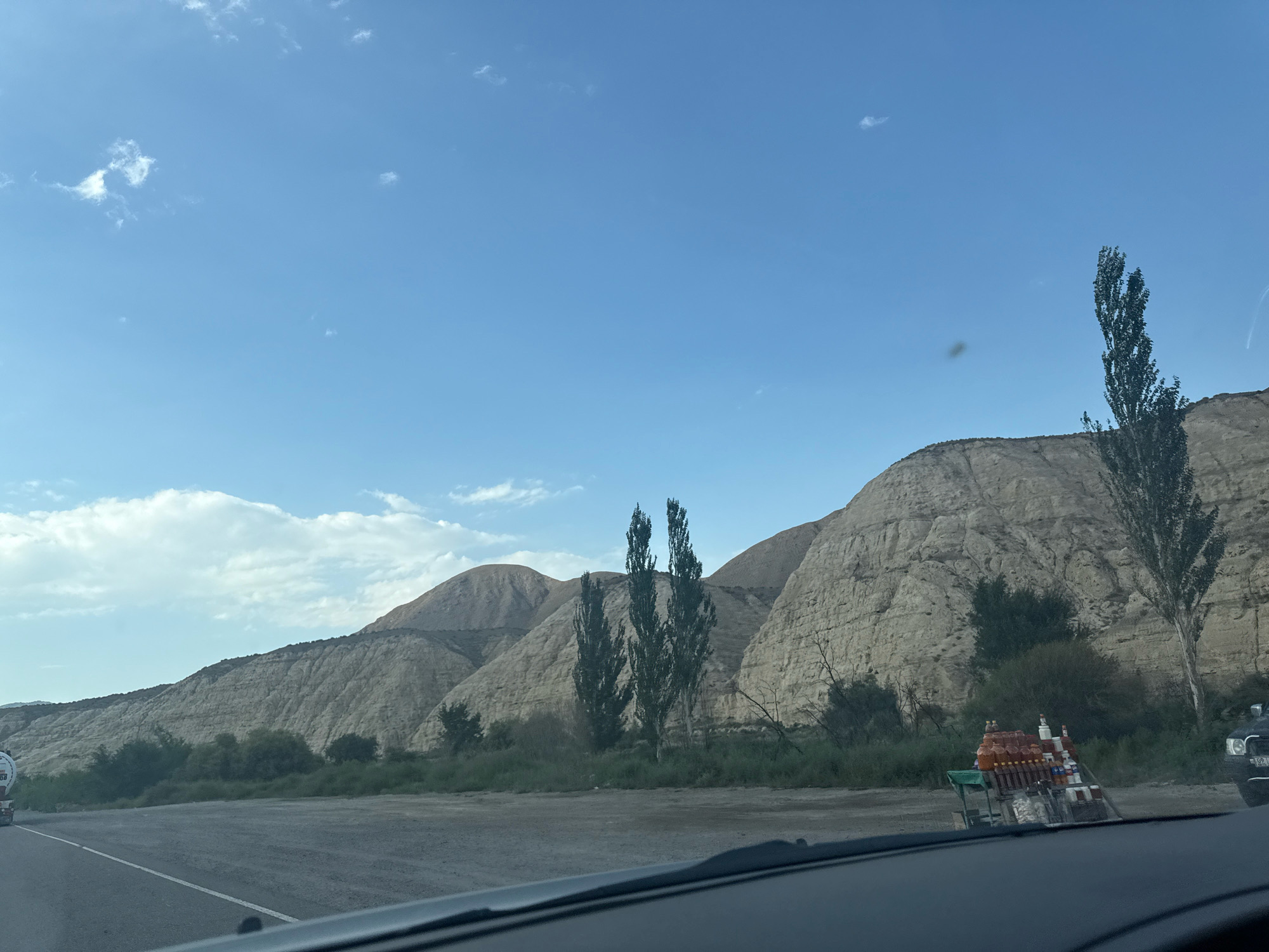 The image depicts a roadside scene in a mountainous, arid region, possibly in Kyrgyzstan, given the mention of a city in Kyrgyzstan. A paved road runs along the bottom left of the frame. In the middle ground, a small cart is laden with numerous bottles of beverages, suggesting it is a vendor's stall. Behind the cart, there is a dense line of green vegetation, with several tall, slender trees interspersed within it. The background is dominated by large, sloping hills and mountains with a dry, layered geological appearance. The sky is a clear blue with a few wispy white clouds. A large truck with a white tank is visible on the far left edge of the road, and the front of a dark-colored vehicle, likely the one from which the photo was taken, is visible at the very bottom of the frame. The overall impression is one of a remote, sun-drenched location along a travel route.