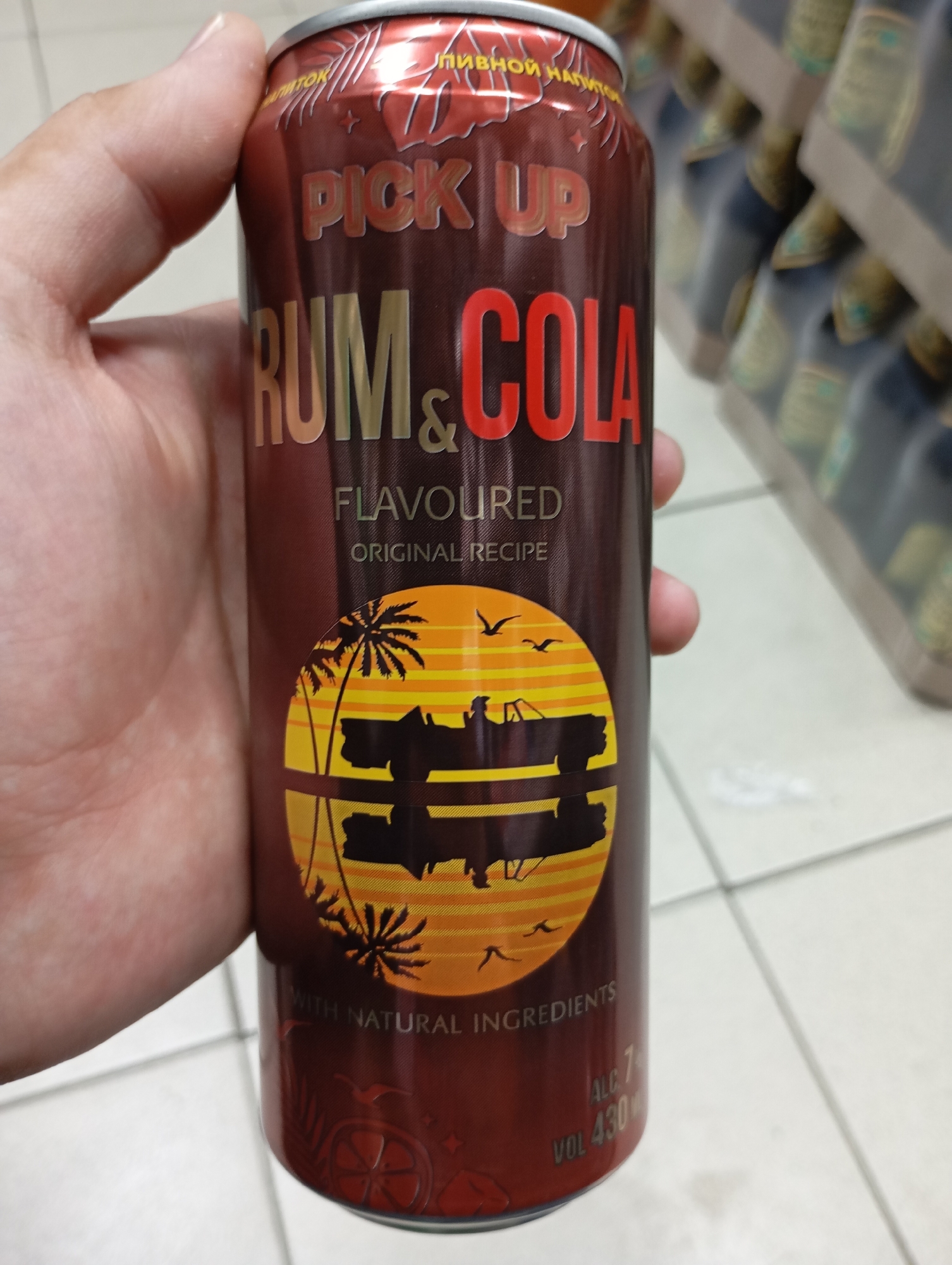 A person's hand is holding a dark red aluminum can of "PICK UP RUM & COLA" flavored beverage. The can has a metallic sheen and features a stylized sunset scene with palm trees and birds. Text on the can includes "FLAVOURED", "ORIGINAL RECIPE", "WITH NATURAL INGREDIENTS", and "ALC 7% VOL 430ml". The background appears to be a store aisle with shelves of similar products blurred out. This image was taken in Moscow, Russia.
