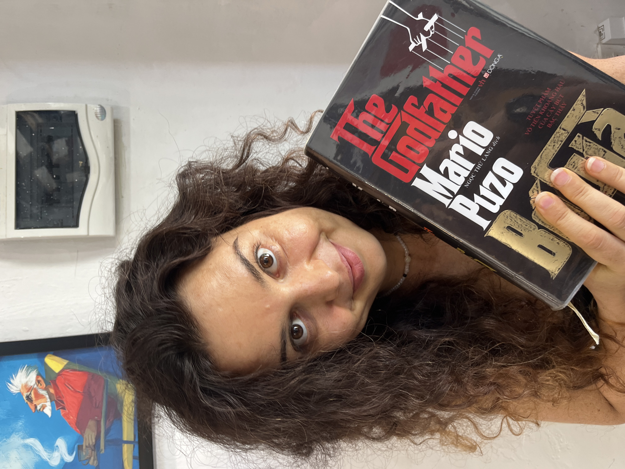 A woman with curly brown hair smiles directly at the camera while holding a book titled "The Godfather" by Mario Puzo. The book is black with red and white lettering, and features a silhouette of a hand manipulating puppets. The woman is wearing a white beaded necklace. Behind her, a framed picture shows an older man with white hair and a beard sitting in a chair and smoking. To the right of the picture is a white electrical panel mounted on a white wall. The overall scene appears to be indoors, possibly in a home or office. There is no visible indication of the location being specifically in Phu Thuy Ward, Vietnam; the environment is generic for an indoor setting.