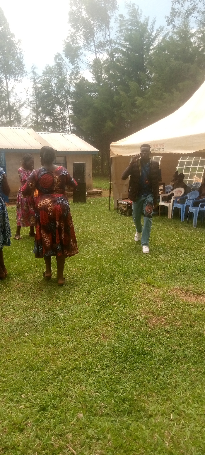 The image shows a scene in a grassy outdoor area, likely a rural setting. Several people are visible. Two women are dressed in brightly colored dresses with floral patterns. One woman is facing the camera, and the other is walking away from the camera. A man, dressed in a brown jacket, blue jeans with a tear in the knee, and white sneakers, is walking toward the camera. He appears to be speaking on a mobile phone. In the background, a large, white tent is partially visible. Several blue chairs are situated around the tent. The image was taken in Kivaywa, Kenya, at the coordinates (latitude: 0.56, longitude: 34.806).