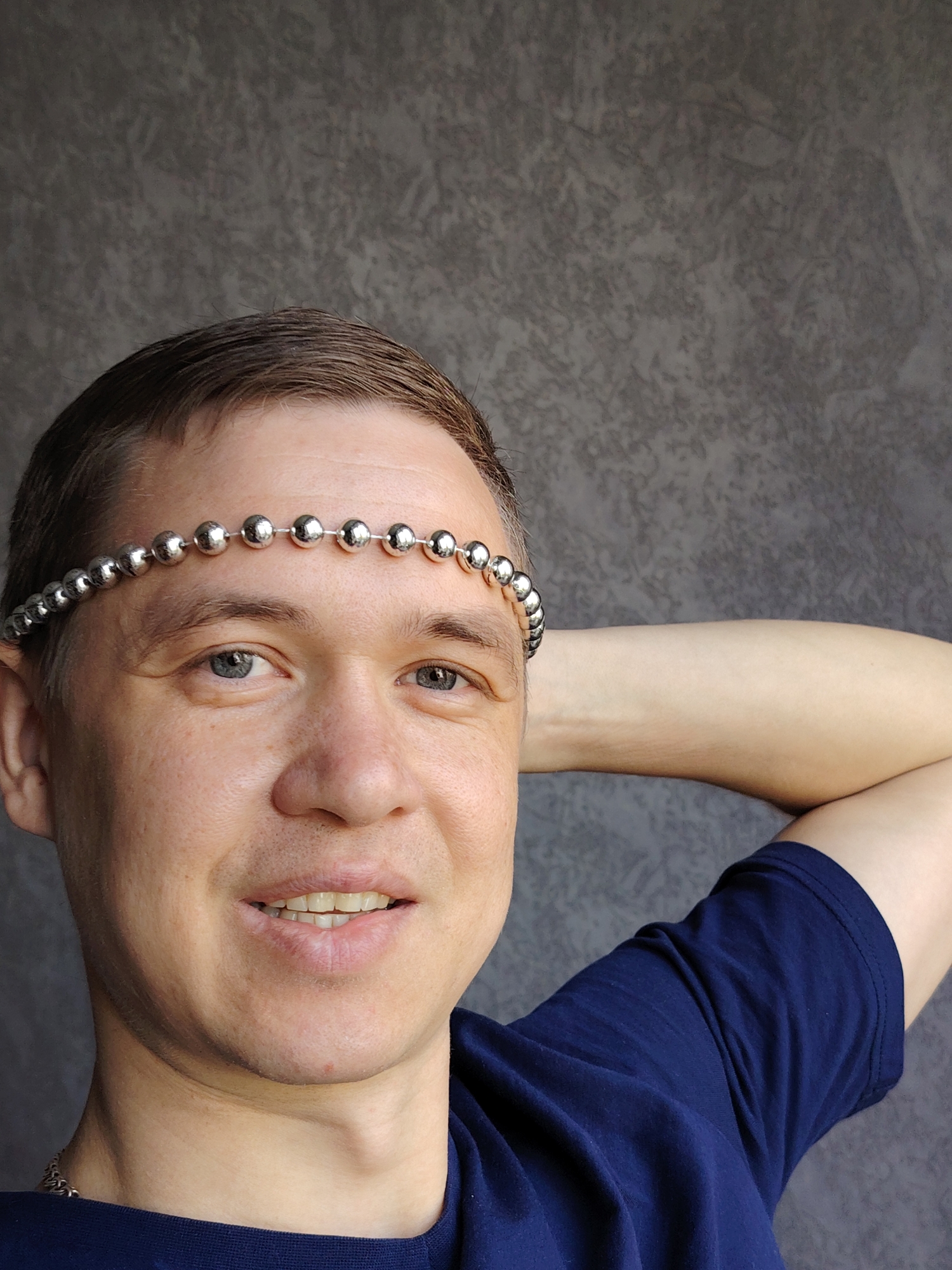 A waist-up portrait photograph features a man with short brown hair, appearing to be in his 30s or 40s, looking directly at the camera with a gentle smile. He is positioned against a dark gray, textured wall, suggesting an indoor setting. His blue-gray eyes are open and engaged. A silver-colored beaded headband or chain is worn across his forehead. He is dressed in a dark blue, short-sleeved t-shirt, and a silver-colored chain or necklace is partially visible around his neck on the left side of the frame. His right arm is bent, with his hand resting behind his head or neck. The lighting is soft and even, highlighting his facial features and the metallic sheen of the beads. The man's expression conveys a pleasant and relaxed demeanor. The location context for this image is Chaikovsky, Russia. There is no visible text in the image.