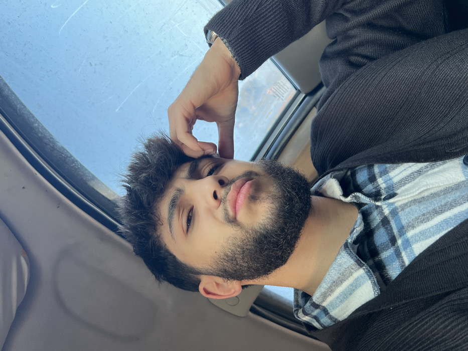 A young man with a beard poses in what appears to be a car interior. He wears a black jacket over a blue and white checkered shirt. His hand is near his forehead, possibly resting or posing. A window is visible in the background, seemingly showing a cloudy or rainy sky. The photo is taken at an angle, adding a dynamic element to the composition.