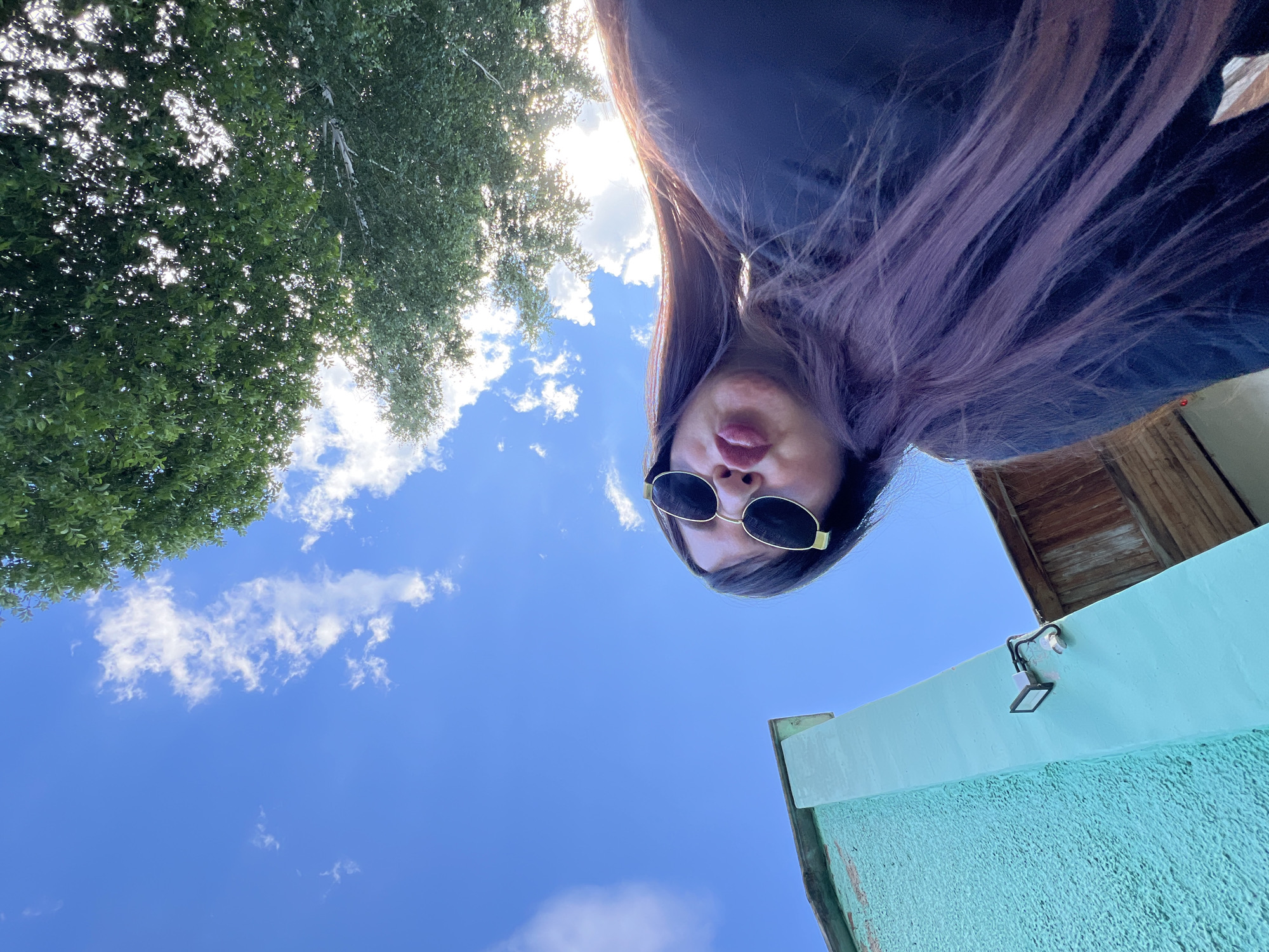 The media file shows a low-angle, wide shot of a young woman with long, purplish-brown hair and sunglasses, looking up towards the sky. She is outdoors, with a bright blue sky and scattered white clouds visible. To the left, the lush green foliage of a tree is seen, with sunlight filtering through its leaves. The woman is making a pouting or kissing face. In the background, to the right, a portion of a building is visible, constructed with wooden planks at the top and a turquoise-colored textured wall below. A small solar-powered light fixture is attached to the turquoise structure. The overall impression is a casual, outdoor selfie taken on a sunny day. No visible text is present. The location context is Noviy, Russia.