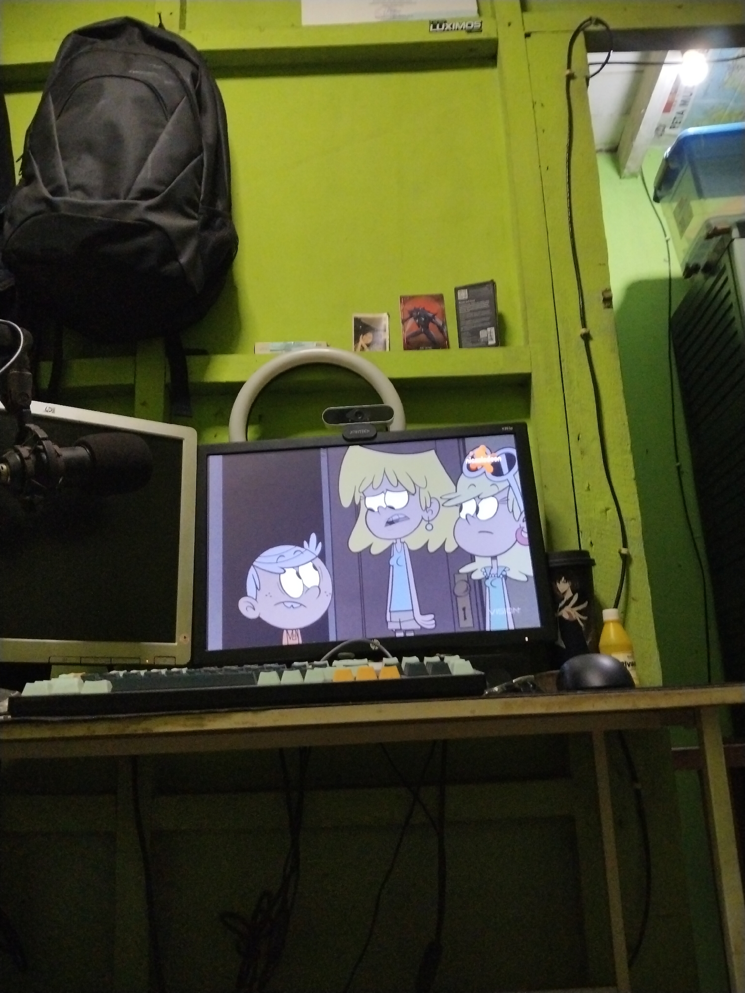 The image depicts a desk with two computer monitors, a keyboard, a microphone, and a backpack hanging on a green wall. The monitor on the right displays an animated scene from "The Loud House" on Nickelodeon, featuring characters Luan and Lori Loud. A ring light is positioned above the monitors, and a webcam is attached to the right monitor. On the wall above the monitors are several books or boxes. The desk is made of wood, and cables are visible hanging underneath. A yellow bottle and a black mouse are also on the desk. The environment appears to be an indoor setting, possibly a room. The overall impression is that of a home office or gaming setup.