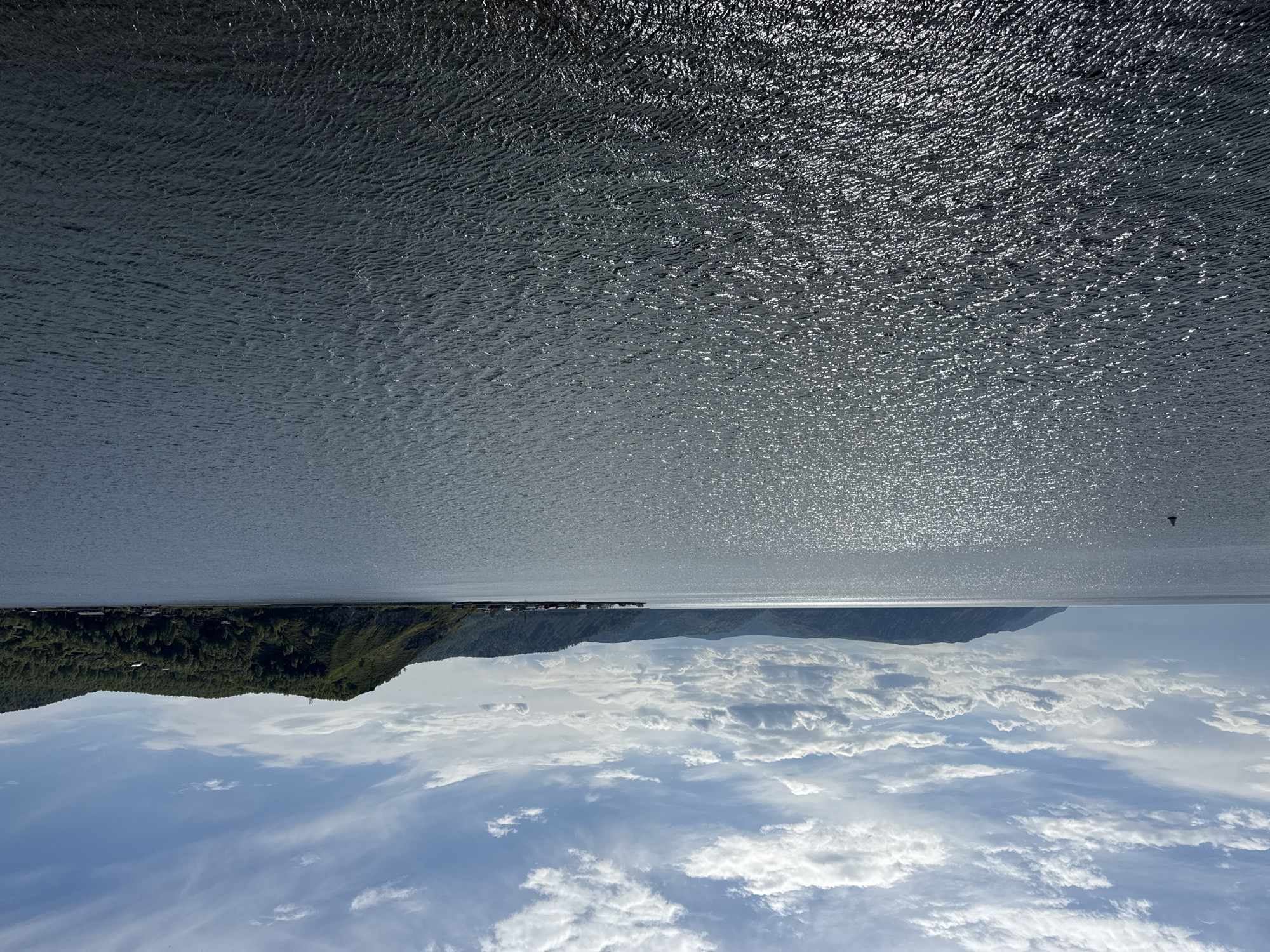 The image shows a landscape in Listvyanka, Russia, oriented with the sky at the bottom and the water at the top. The sky is blue with many white clouds. Above the sky is a landmass with green trees and some buildings along the shore. The land appears to be connected to a distant mountain range. Above the land and mountains is a body of water, with a glistening, textured surface that reflects light. The image is taken from an elevated point, perhaps from a moving vehicle.