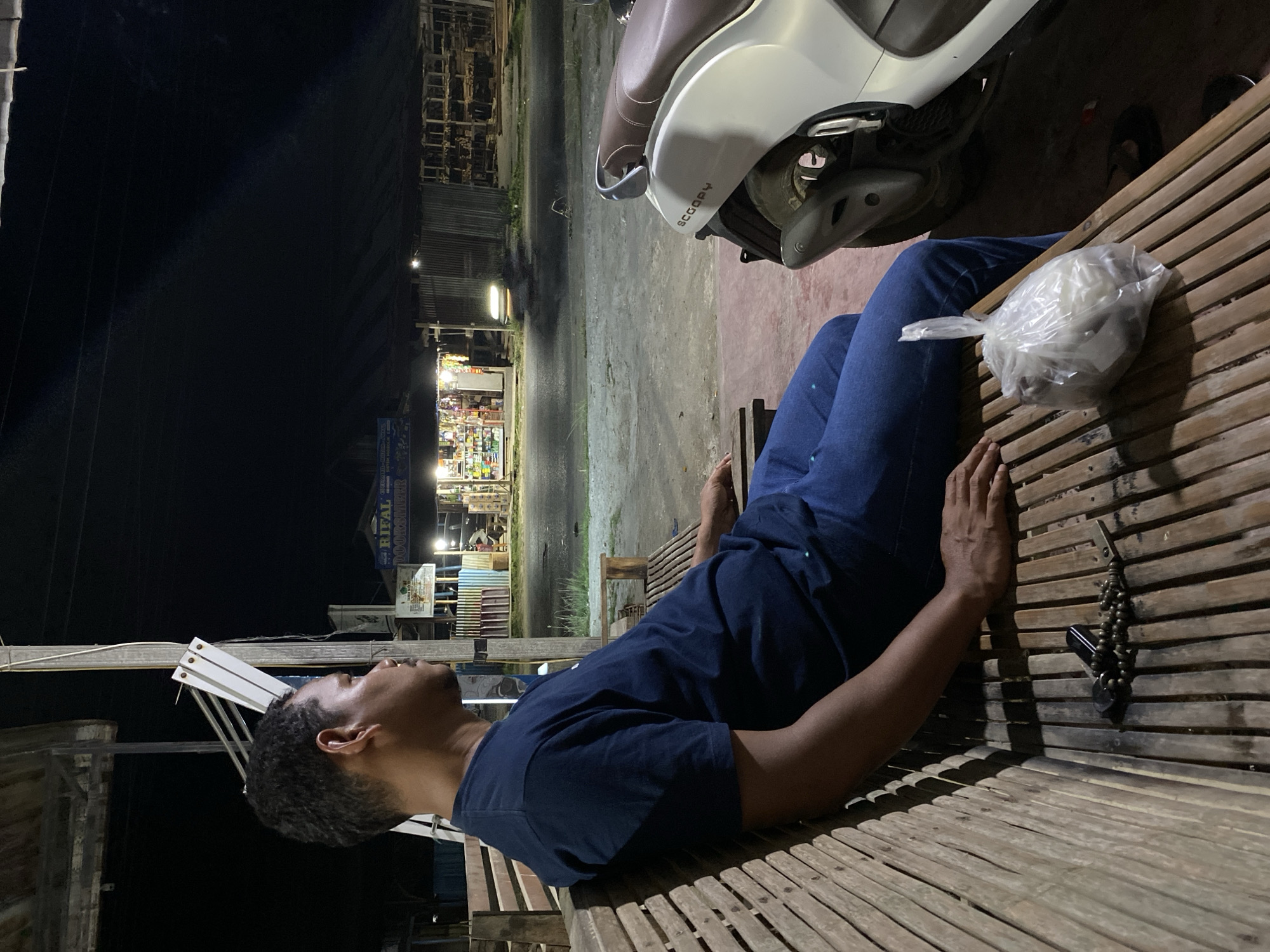 A man is lying on a wooden bench in Teupinbatee, Indonesia, adjacent to a scooter and a store. He is dressed in a dark blue t-shirt and blue jeans. His keys with a beaded chain lie next to him on the bench. A plastic bag is resting on his leg. The setting is at night; the street is lit by store lights.