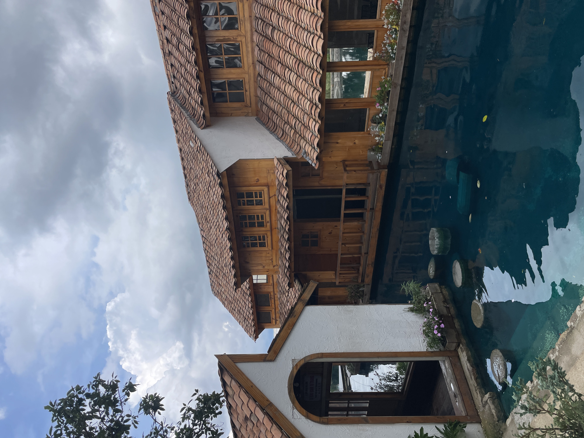 A wooden building with terracotta roofs stands beside a body of turquoise water. The building features multiple windows and balconies, and a smaller white structure with an arched doorway is attached to its left. Several stepping stones are submerged in the water, and some small flowers are planted in wooden boxes along the edge of the building. The sky above is partly cloudy. This scene is located in Thôn 3, Vietnam.