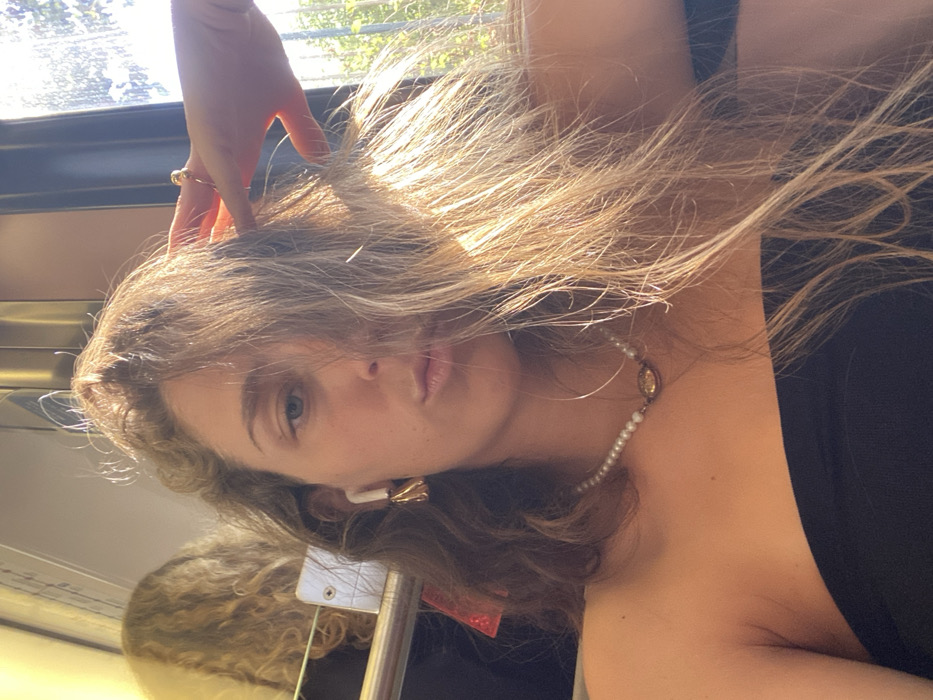 The image depicts an indoor portrait of a female subject, likely a selfie, taken within a vehicle in Kallithea, Greece. The subject, positioned centrally, has long, wavy hair with light brown and blonde tones, which partially obscures the left side of her face. Her visible right eye is blue, and her lips are pursed. She wears a black strapless top and a white pearl necklace featuring a central oval, gold-toned pendant. In her right ear, a white wireless earbud is visible, accompanied by a gold-toned, irregularly shaped dangling earring. Her right hand, adorned with a gold-toned ring on the index finger, is raised and positioned within her hair. Strong natural light illuminates her from the right. The background on the right features a window showcasing blurred green foliage outside. To the left, a reflection reveals the partial head of another individual with light, curly hair, alongside interior vehicle elements including a silver-toned horizontal bar and a white rectangular fixture.