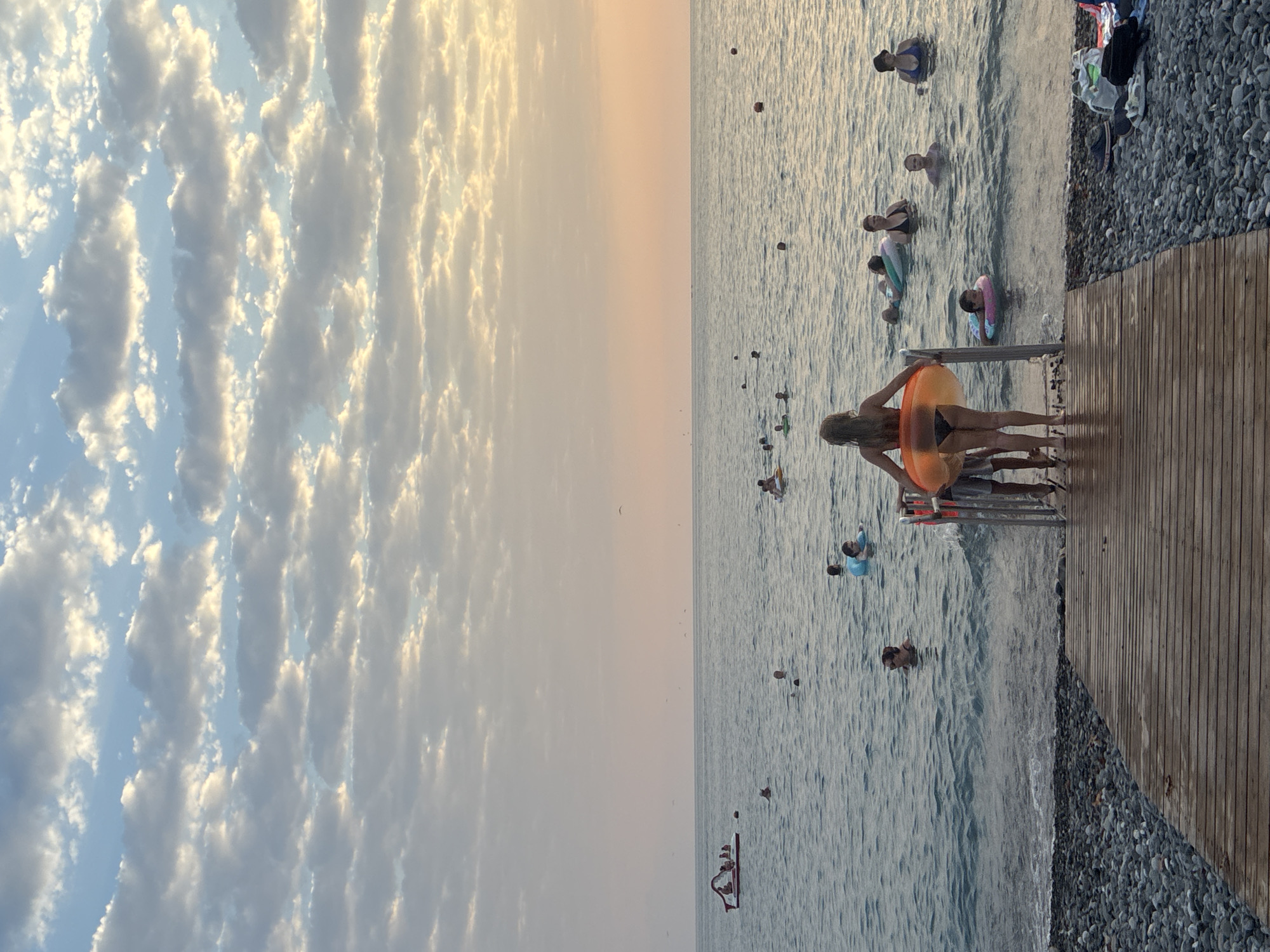 An outdoor, eye-level, full shot captures a wide view of the sea at sunset, with a wooden pier in the foreground and a beach of smooth grey stones to the right. A group of people are visible in the water, some swimming and others on inflatable devices. The sky is filled with textured clouds illuminated by the setting sun, casting a warm glow over the scene. The location is a beach in Sochi, Russia.