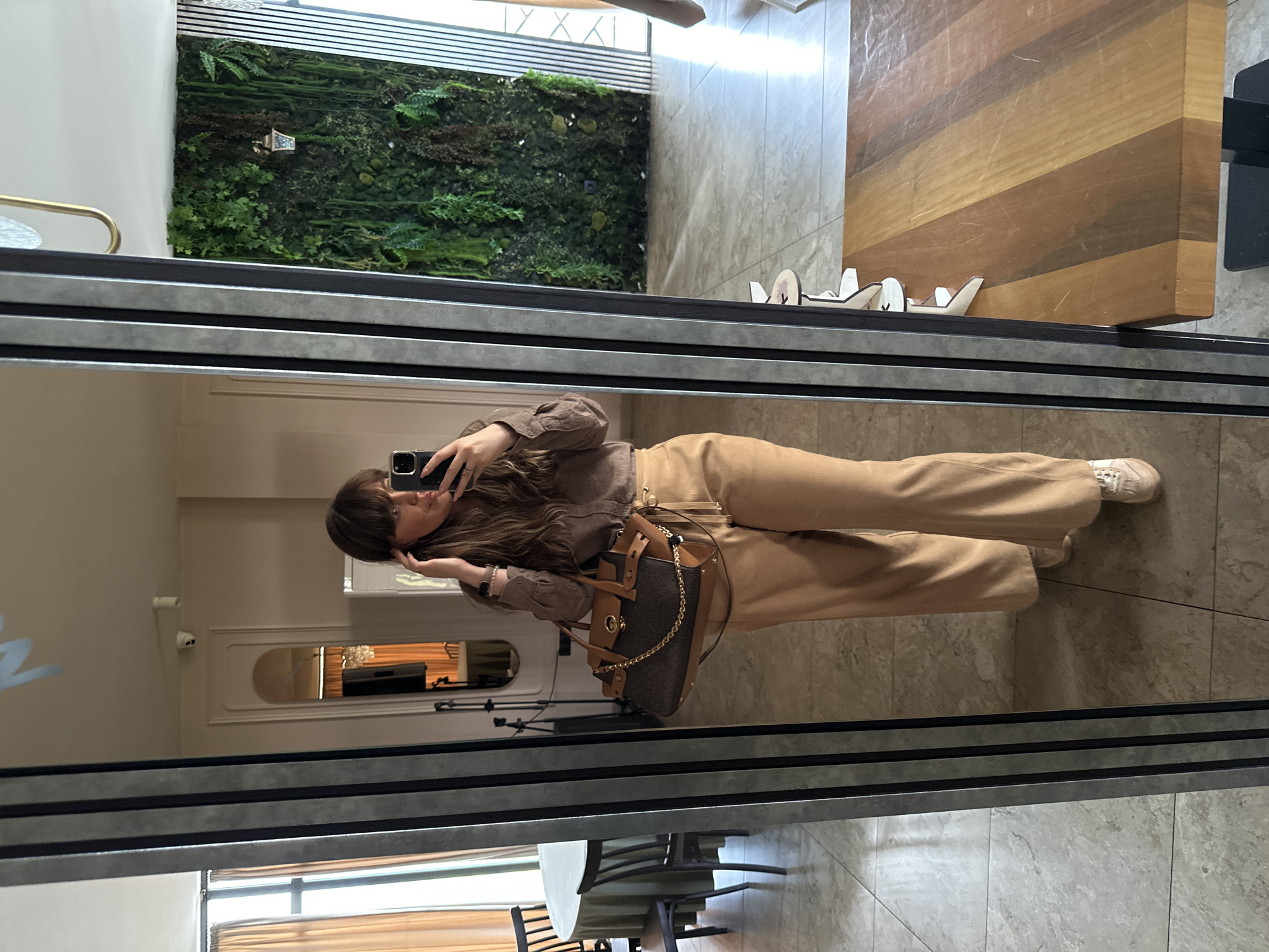 A woman is seen in a full-body reflection in a mirror. She is wearing a brown button-up shirt and beige pants and has a brown handbag hanging from her shoulder. She is holding up a phone, presumably taking a photo of herself in the mirror. The background reflected in the mirror shows a room with a table and chairs, a doorway, and a wall with some blue writing on it. The mirror is in a space that has a tiled floor, a green wall, and a wooden tabletop. Based on the interior design elements, it is possible that this image was taken in a cafe or restaurant. It is not possible to confirm the specific location in Marneuli, Georgia without further information.