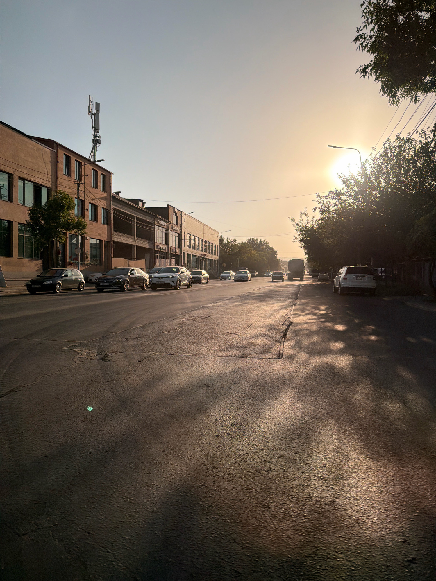 This is a street scene in Yerevan, Armenia, captured during the late afternoon or early evening, evidenced by the low angle of the sun casting long shadows. The sun is low in the sky, partially obscured by trees and power lines, creating a warm, hazy light. The street itself is asphalt with visible cracks and patches. On the left side of the street, there are multi-story buildings with a cell tower mounted on the roof of one. Several cars are parked along the sidewalk or driving on the street. On the right side of the street, there are trees and more buildings. The overall impression is of a somewhat older urban environment with a mix of residential and commercial structures.