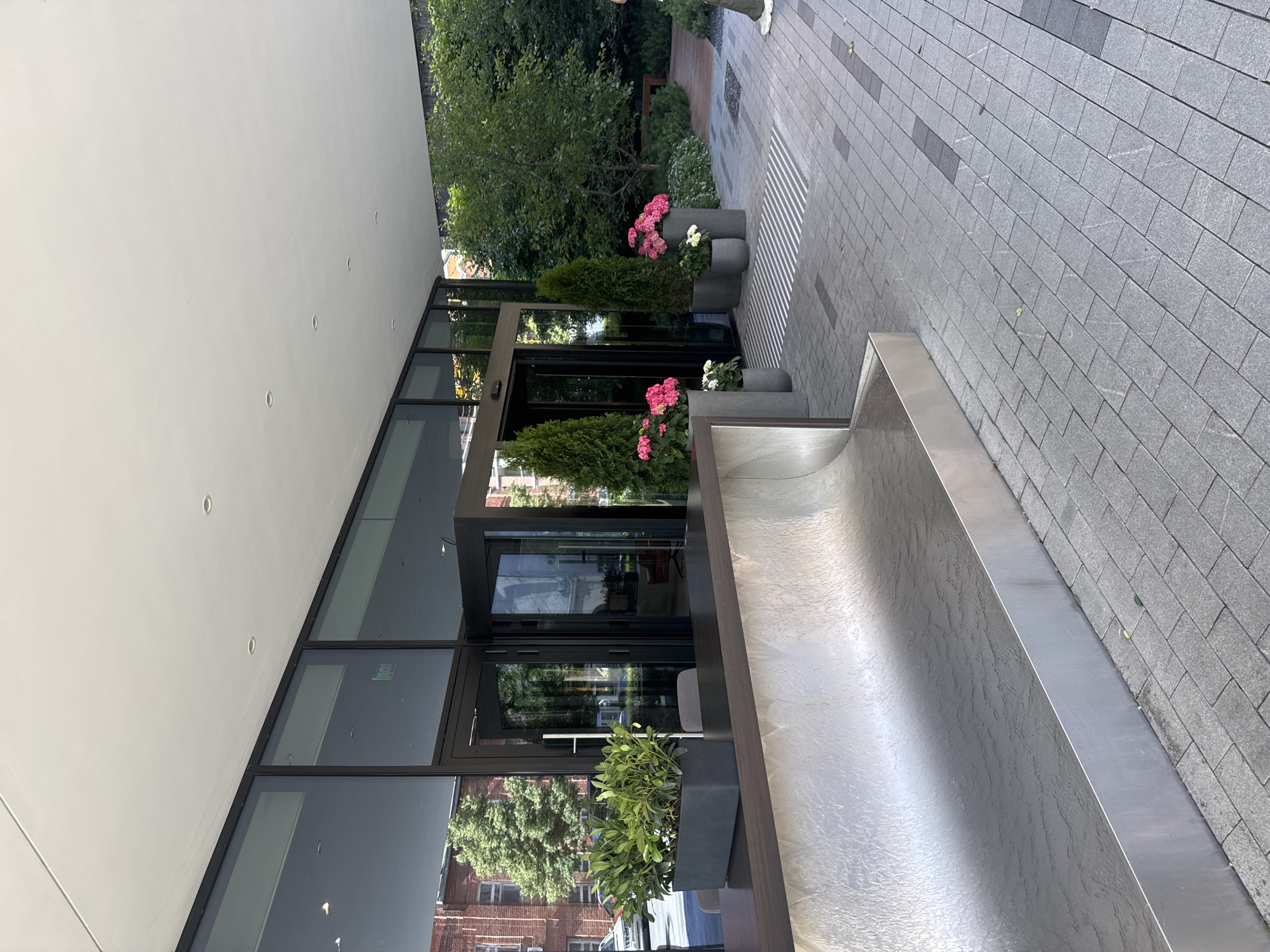 The image shows the entrance to a modern building with large glass doors and windows. In front of the entrance, there is a metal water feature with a textured surface. Several potted plants are placed around the entrance, including tall evergreen trees and flowering bushes with pink and white blossoms. The ground is paved with gray rectangular stones, and there are tactile paving strips for visually impaired pedestrians. In the background, trees and foliage suggest a green outdoor space. To the left, a building with red brick facade is visible through the glass. The scene appears to be a public space, possibly a commercial or residential building entrance in Yekaterinburg, Russia.