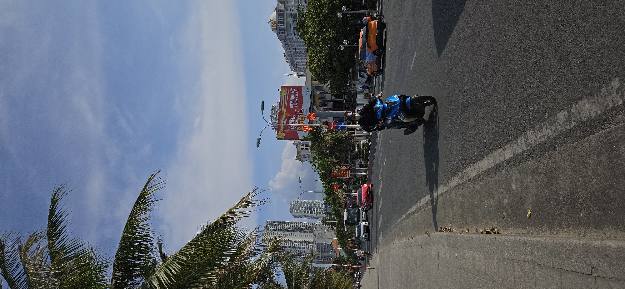 This image depicts a street scene in Nha Trang, Vietnam, characterized by urban infrastructure and tropical vegetation. A person on a blue motorcycle drives on the right side of the road, away from the viewer. An orange three-wheeled vehicle is in the opposite lane.
The road is paved, with a painted white line separating traffic lanes. Debris is scattered along the curb. Palm trees line the left side of the road, with buildings visible in the background. A billboard displays Vietnamese script. The sky is predominantly blue with scattered clouds.