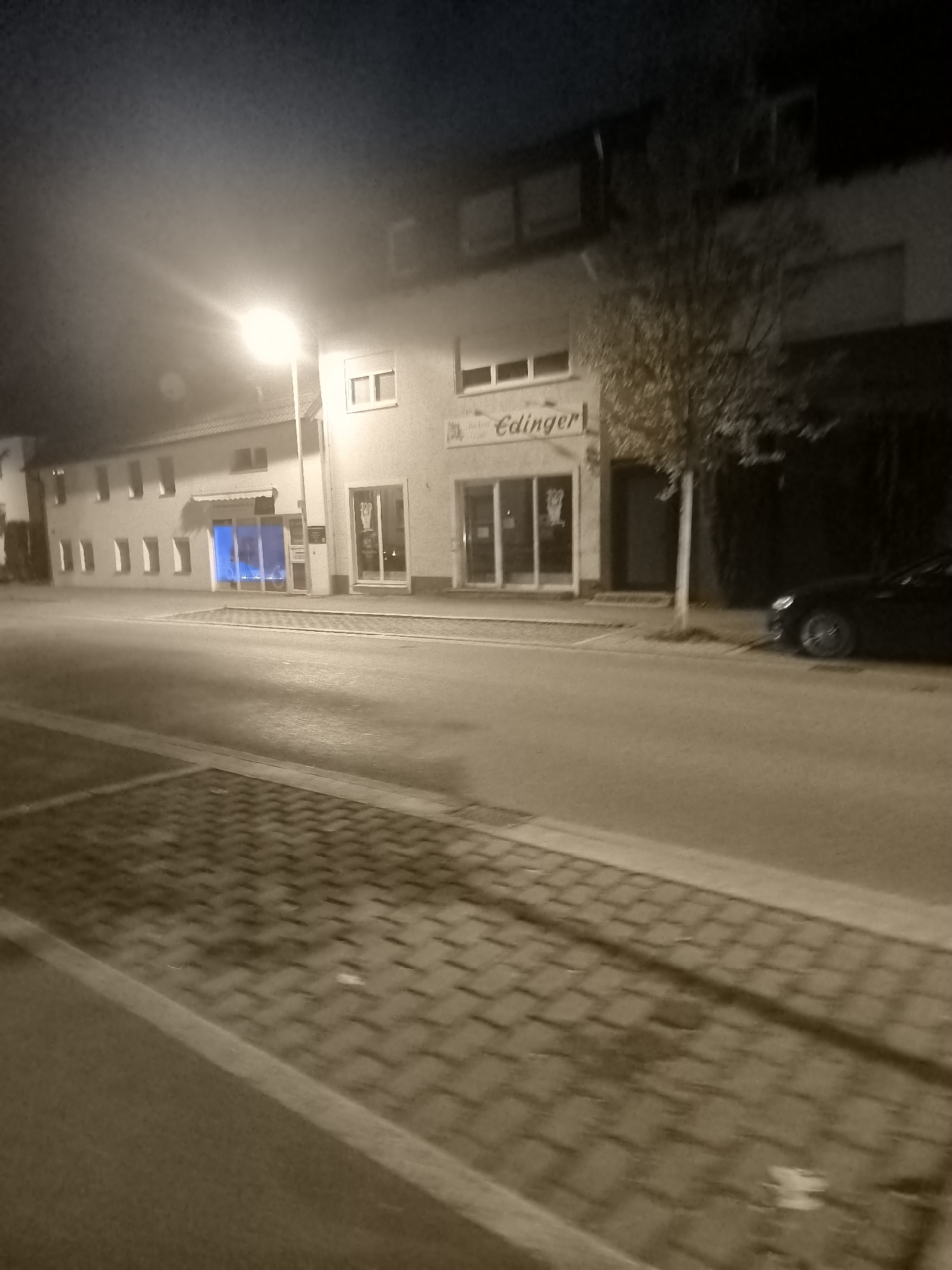 A street scene at night in an unknown city. A streetlight illuminates a section of a building with multiple windows and an awning over one of the entrances. To the right of this building is a darker building, and next to that is a tree. A black car is parked on the street in front of the darker building. The foreground shows a sidewalk paved with interlocking bricks. The overall image has a sepia tone, suggesting it was taken with low light or a filter.