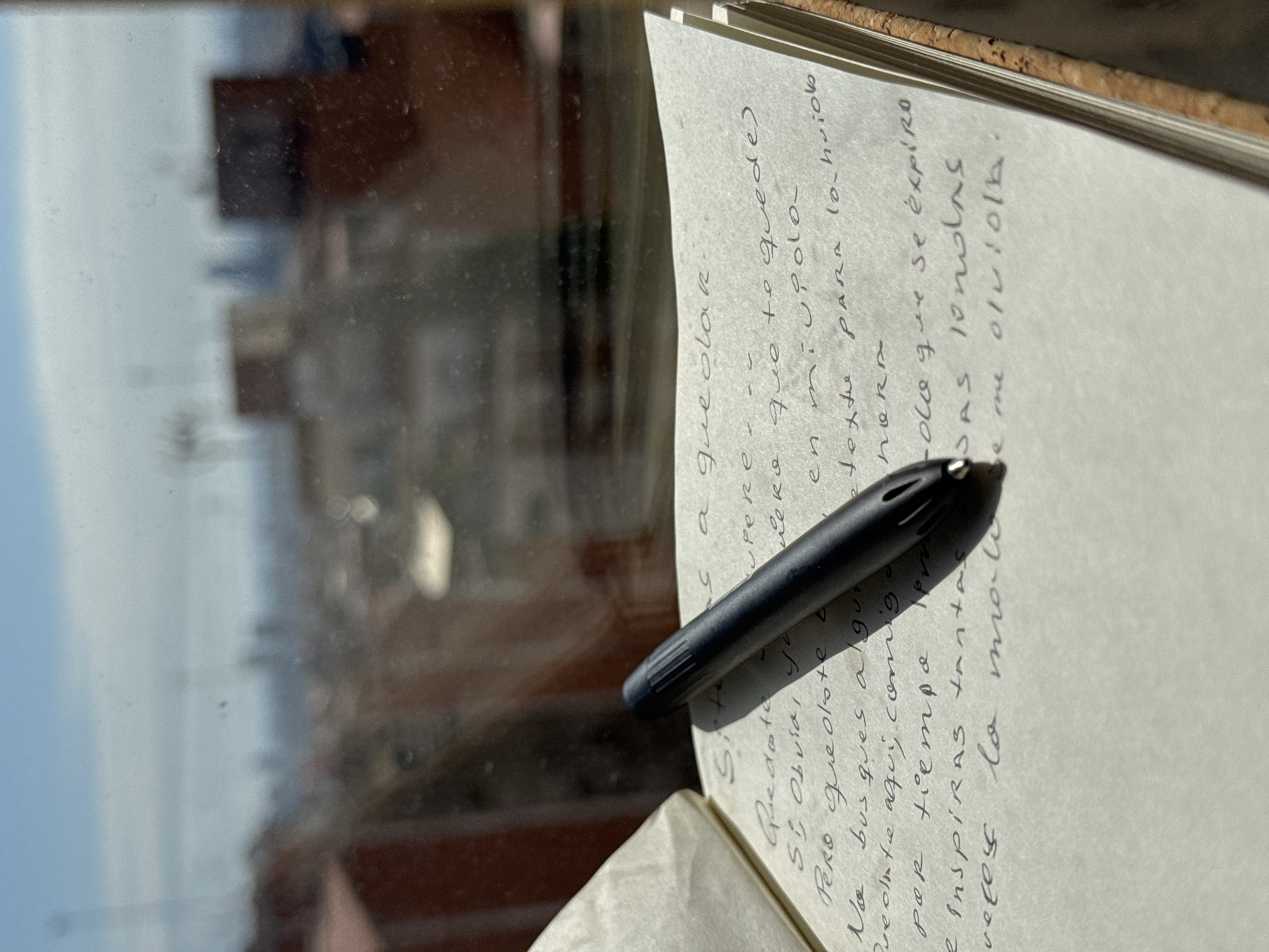 The image depicts a notebook with handwritten text in Spanish and a black pen resting on it. The notebook is open, and the pages are slightly yellowed. The text appears to be a poem or a personal journal entry. A black pen with decorative cut-outs near the tip lies on the page, partially obscuring the writing. Through the window is a blurred cityscape with buildings under a bright, hazy sky. The exact location within Barcelona is not discernible from the image.