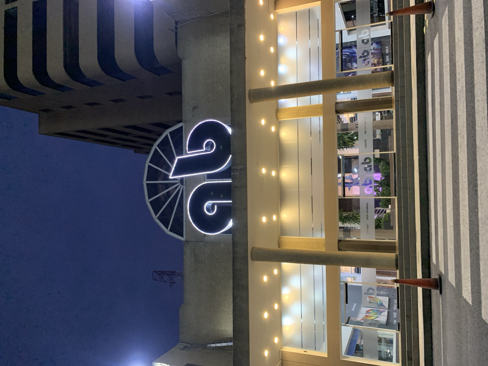 The image, originally rotated 90 degrees clockwise, captures an urban night scene featuring a modern building facade and a prominent illuminated sign.
The main building, situated on the right side of the correctly oriented image, displays a sleek, multi-story facade composed of glass panels and light-colored vertical elements. Numerous small, warm-toned lights are embedded within these vertical sections, casting a glow across the structure. Two substantial concrete columns extend horizontally across the middle section of the facade, possibly part of a covered entrance or architectural feature. Reflections on the glass offer glimpses into the illuminated interior, revealing what appear to be commercial or office spaces.
To the left of this building, a large, brightly glowing white neon sign is affixed to a grey concrete wall or pillar. The sign clearly depicts the number "26", stylized with a distinct, flowing design. Above and to the left of this sign, against a dark blue night sky, a portion of another building with distinctive curved, tiered architectural elements is visible. A faint, skeletal metal structure, possibly a crane or antenna, can also be discerned in the distance against the dark sky.
In the foreground, on the right side, a pedestrian zebra crossing with alternating white and grey stripes is visible. Two orange traffic cones are positioned near the crossing, indicating a temporary boundary or warning.
The scene is set at night, evident from the dark blue sky and the pervasive artificial lighting from the building and the illuminated sign. No people or specific activities are visible, contributing to a serene urban atmosphere. Visible text on the glass panels of the main building includes "CENTRO EMPRESARIAL" and a repeating, often mirrored, "gb" logo or lettering. The overall setting suggests a commercial area in Pampatar, Venezuela.