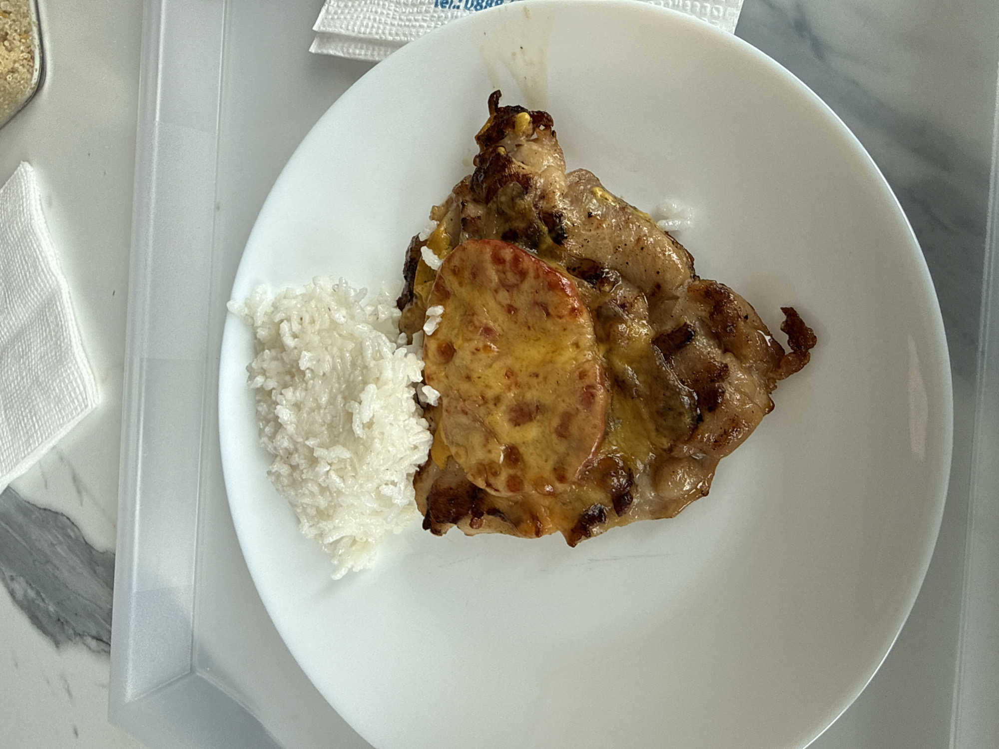 A plate of food is on a white plate. The food consists of a portion of white rice and a piece of meat topped with a slice of sausage. The plate is placed on a white surface next to a container and some napkins. The image was taken in Sofia, Bulgaria.