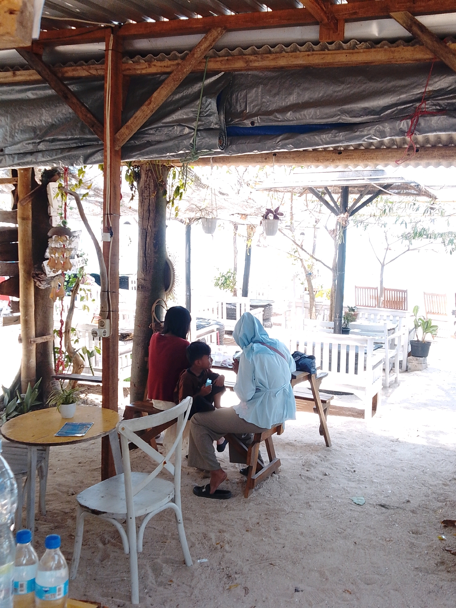 This is a medium shot photograph taken at what appears to be an outdoor cafe in Cikal, Indonesia. The scene is set under a rustic, partially covered structure supported by wooden beams. A grey tarp is visible overhead, creating shade. The ground is sandy, characteristic of a beach or coastal area.
In the foreground, a round yellow table with a white base is positioned to the left. A small potted plant sits on the table, next to a blue booklet. A white wooden chair with an 'X' back design is in front of the table, facing away from the camera. Two plastic water bottles are on the table in the lower left corner of the frame.
The central focus of the image is a group of three people sitting at a wooden table. A person wearing a red top is seated on the left, with their back to the camera. Next to them, a young boy in a brown shirt is holding a smartphone. To the right of the boy, a person wearing a light blue hooded outfit, possibly a hijab, is also seated. They are all facing away from the camera and seem to be engaged with each other or the items on the table.
Behind the central group, the cafe extends with more white wooden seating and tables, some of which are covered in shade. Several potted plants are scattered around the seating area. In the background, trees and foliage are visible, suggesting a natural environment surrounding the cafe. The lighting indicates it is daytime, with bright sunlight filtering through the overhead cover.