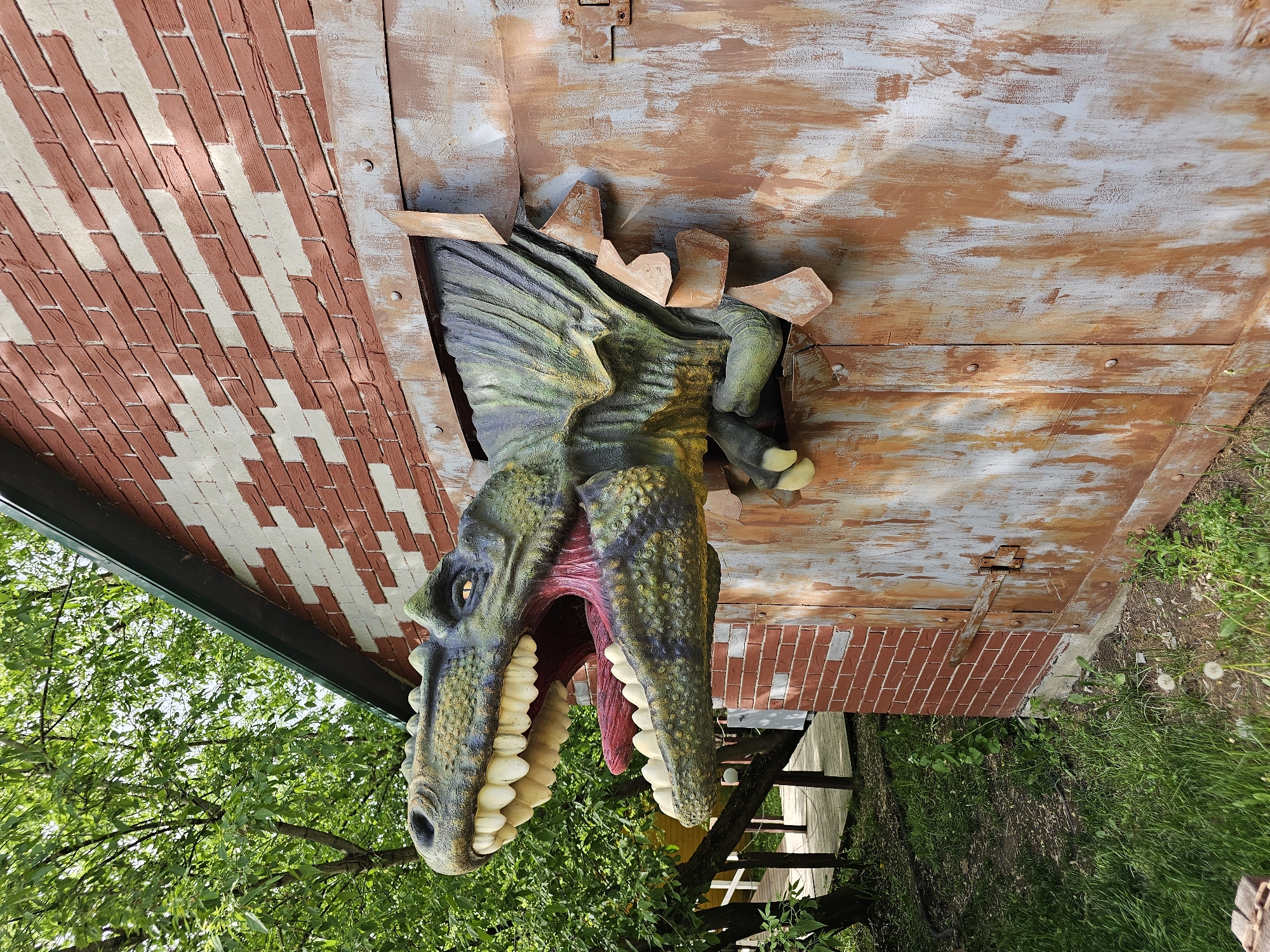 A dinosaur head protrudes from a hole in the side of a building. The building has sections painted to resemble bricks and weathered metal siding. The dinosaur is green and yellow with large white teeth and a red tongue. Trees and foliage surround the building. The image appears to be taken outdoors. There is no evidence to verify its exact location in Khimki, Russia.