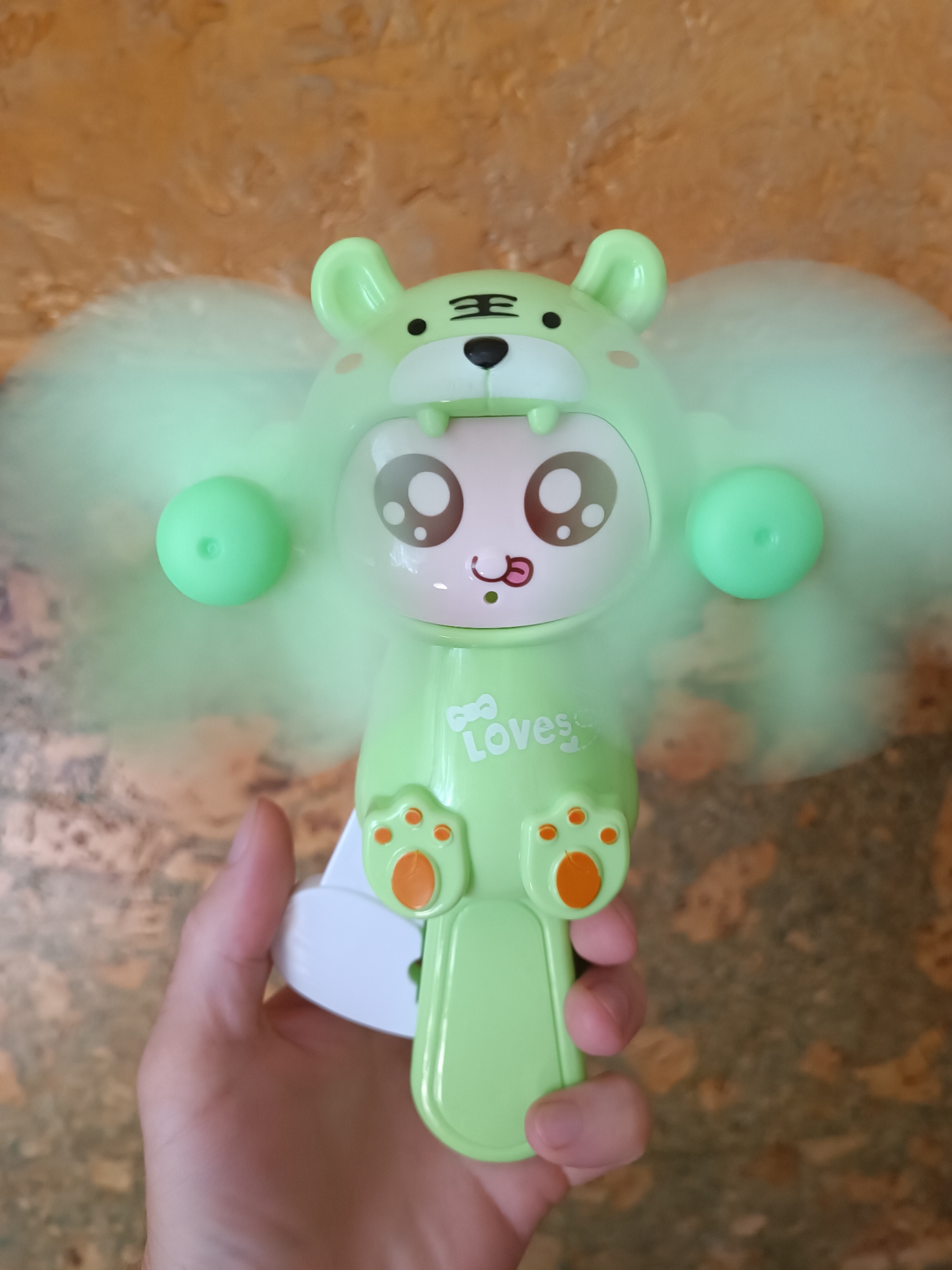 A light green plastic toy fan, shaped like a cartoon tiger face with large eyes and ears, is being held by a person's hand. The fan blades, also light green, are spinning and create a blurred effect. The tiger face has black stripes on its forehead, small black eyes, and pink cheeks. Below the face, the word "Loves" is visible in white text, followed by paw prints. The background is an abstract textured surface, appearing to be a wall or floor. The scene is indoors, in an environment that is not specified as being in a city or having a specific location.