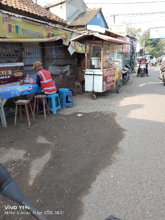 The image shows a street scene in Kecamatan Haurgeulis, Indonesia. A man wearing an orange vest is sitting at a small, blue and white table. He is seated on a blue stool, and two red stools are near him. The table is covered with a blue tablecloth. There is a food cart in the background. There are other people in the background, but they are not in focus. The street is paved and there is a small amount of dirt on the left side. A motorcycle is parked in the background. The sky is bright.