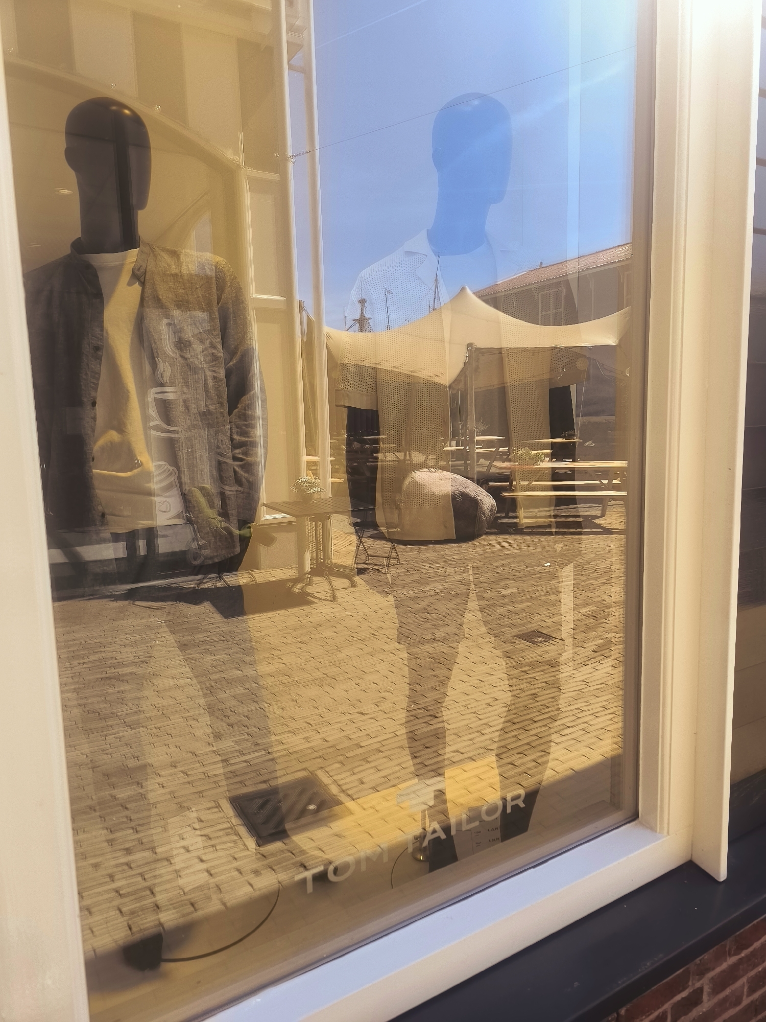 This image shows a reflection in a storefront window, likely located in Lelystad, Netherlands, given the presence of "TOM TAILOR" branding, a well-known clothing retailer with stores in the Netherlands. The reflection reveals a bright, sunny outdoor scene.

In the foreground of the reflection, a mannequin is visible inside the store, wearing a grey, textured jacket over a light-colored shirt. The store's interior appears to be set up with a paved walkway and possibly outdoor seating area, suggested by picnic tables and a large, round beanbag-like seat under a white tent structure.

The reflection also shows the blue sky and part of a building with a tiled roof, indicating an urban or commercial setting. The pavement outside the store is made of light-colored bricks. The text "TOM TAILOR" is printed on the window, indicating the name of the establishment. The overall lighting suggests it is daytime, with strong sunlight casting shadows.