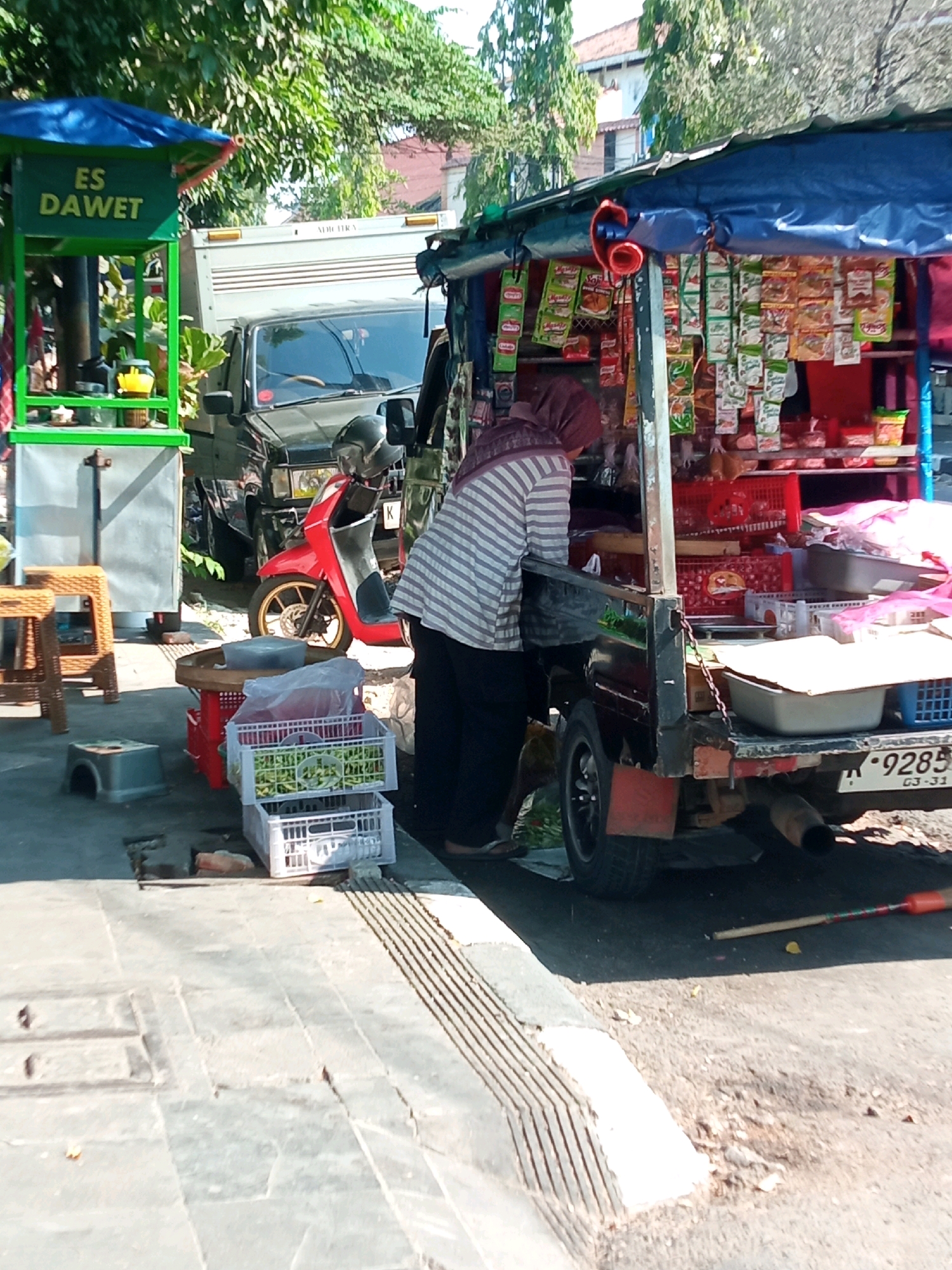 An outdoor scene in Balun, Indonesia depicts a street-side vending operation. In the foreground, a grey sidewalk with textured grates leads to a paved area. To the left, a green food stall with blue roofing displays the text "ES DAWET" in yellow lettering. Beside it, a grey metal cooler and two brown woven stools are visible. A red scooter is parked in front of a black truck.
The main focus of the image is a mobile food cart, constructed from the back of a dark-colored pickup truck. The cart is outfitted with a blue tarp roof and shelves laden with various packaged goods, likely snacks and condiments. A woman, wearing a hijab and a striped shirt, is attending the cart from the driver's side. In front of the cart, several white crates filled with green produce are stacked.
The background shows trees, buildings with terracotta roofs, and a white truck. The overall environment suggests a warm, sunny day.