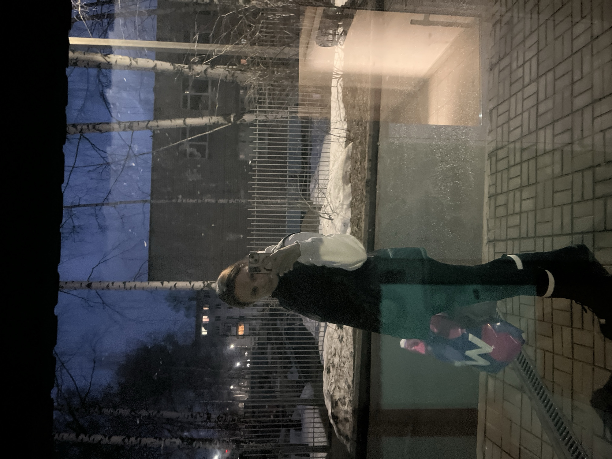 A photograph, likely taken through a window, depicts a young woman reflected in the glass while holding a smartphone and taking a photo. She is wearing a white top, a dark vest, dark pants, and boots with white stripes at the top. The reflection shows a residential street scene at dusk or night. Bare trees, buildings, and a fence are visible. Streetlights illuminate the area. On the other side of the window, a wall composed of rectangular tiles is visible, as well as a drain pipe. The photograph shows details of the girl's clothing and the phone. In front of the girl on the ground, there appears to be a multi-colored item with some visible white patterns. This could be a bag or other object. The location is Chaikovsky, Russia.