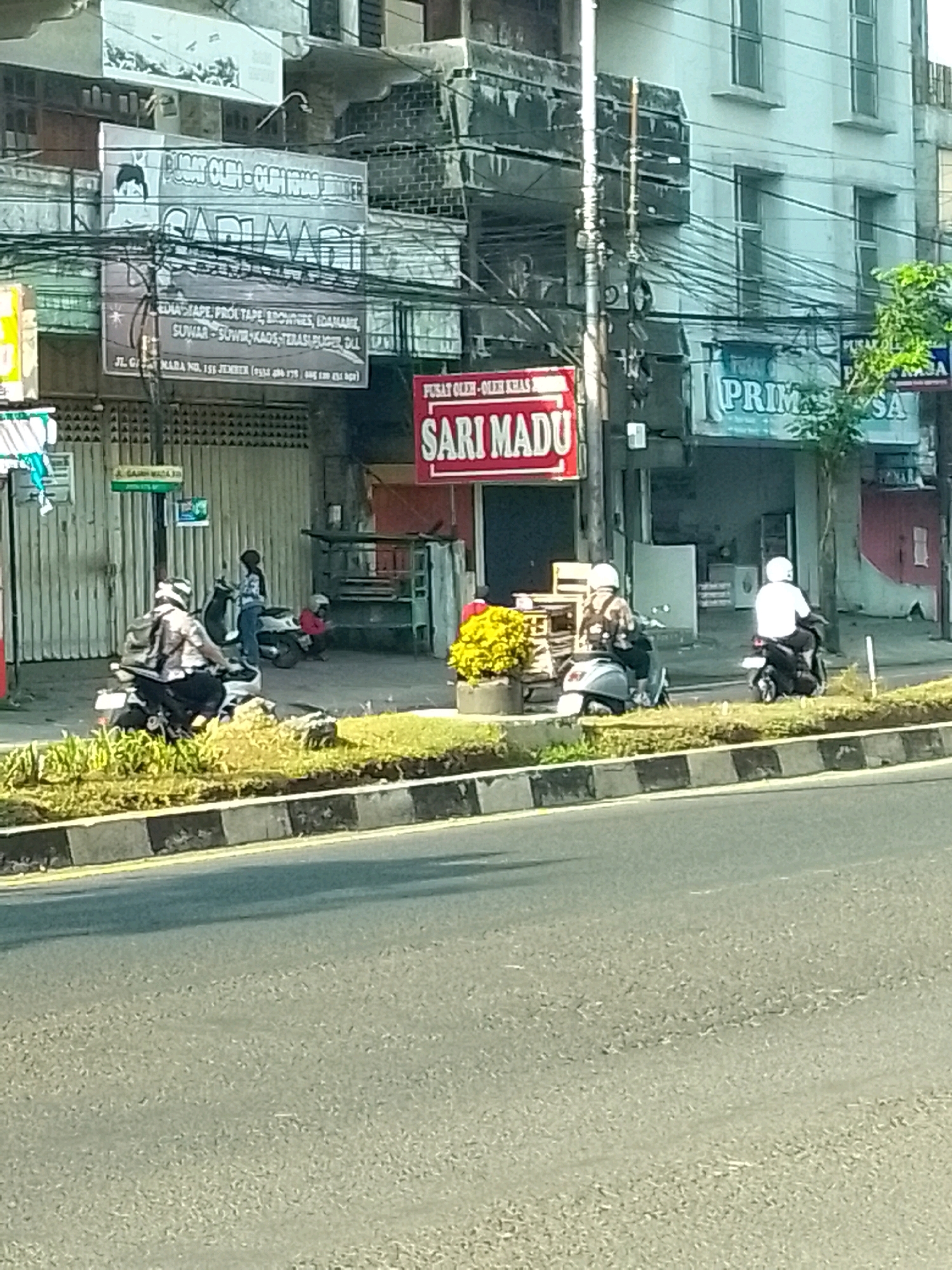 The image shows a street scene in Jember, Indonesia, featuring a row of buildings with various storefronts. In the foreground, a street is visible with a grassy median strip. Several people are on motorcycles. One person on a scooter is wearing a patterned shirt and helmet, parked on the grassy area. Another person on a scooter is wearing a white shirt and helmet, also on the grassy area, facing away from the camera. Further back, a person in a plaid shirt and jeans is standing next to a parked motorcycle.
The storefronts display a variety of signs. One large sign reads "SARI MADU" in red lettering with a white outline, above text describing products and an address on Jl. Gajah Mada, No. 153, Jember. Another sign, partially visible on the right, reads "PRIMA SA". The buildings appear to be two or three stories high, with some showing signs of age and disrepair. Electrical wires are visible strung across the buildings and poles. Yellow flowers are planted in a pot near the "SARI MADU" storefront. The overall impression is a bustling urban commercial area in Indonesia.