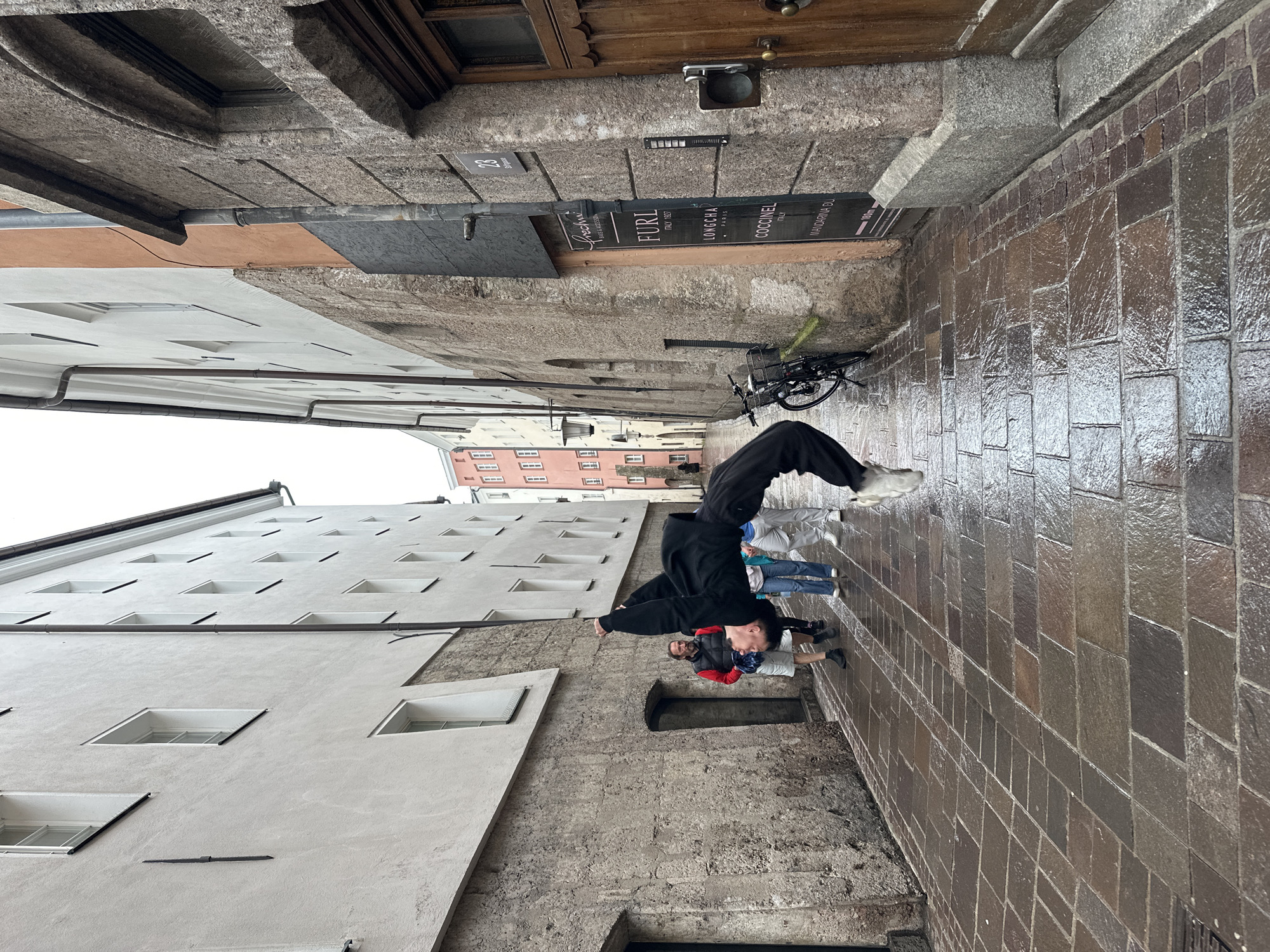 A person is performing a backflip in a narrow cobblestone alley in Innsbruck, Austria. The person is wearing a black hooded jacket and black pants. They are in mid-air, upside down, with their arms extended. Several other people are walking in the alley behind them, some looking at the backflip. The buildings on either side of the alley are old stone buildings with multiple windows. The cobblestones are wet, indicating it may have rained recently. A bicycle is parked against one of the buildings. A sign on the building reads "FUR... ITALY 1927". The sky is overcast.