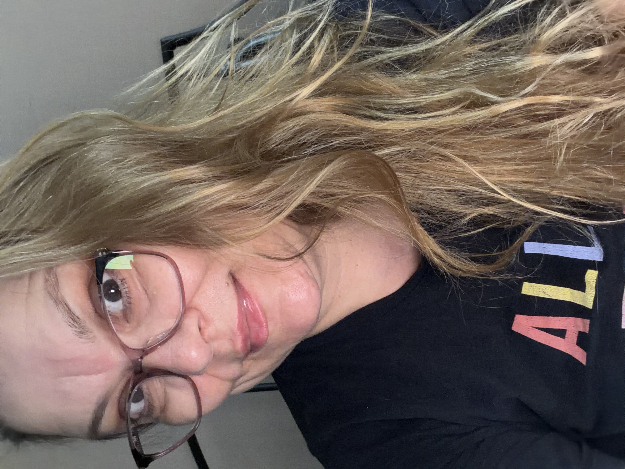 A woman wearing glasses and a black t-shirt is shown from the chest up. The t-shirt has colorful text that reads "ALI". She has long, wavy blonde hair. The background is a plain gray wall. The image is a selfie, taken indoors. No specific location in Southend-on-Sea, United Kingdom can be identified.
