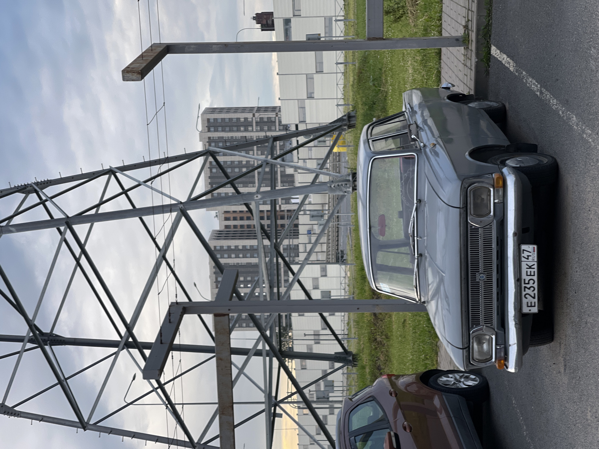 A grey, vintage sedan is parked in an outdoor paved lot next to a brown SUV. The sedan has a license plate that reads "E235EK 47 RUS". In the background, there is a large metal power line tower, a white building with many windows, and several modern apartment buildings under a cloudy sky. The area is grassy and appears to be a public space.