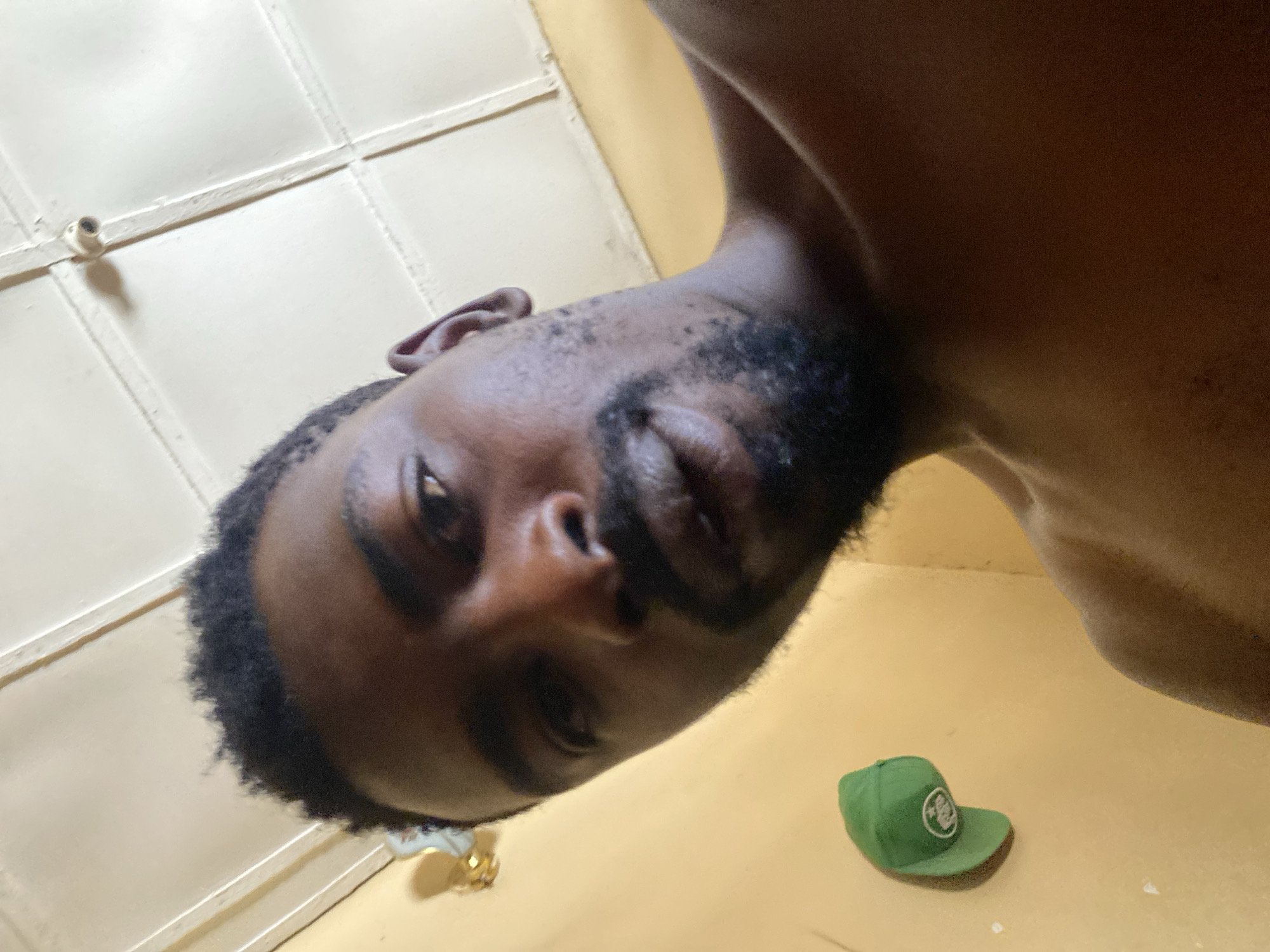 This is a low-angle, close-up indoor selfie of a man, rotated approximately 90 degrees counter-clockwise. The subject is a man with dark skin, a short dark beard, and closely cropped black hair on the sides. He looks directly at the camera with a neutral expression. He appears shirtless, with his shoulder and chest visible towards the right side of the frame.
The background primarily consists of a warm, light yellow or beige wall or floor surface. Above the man's head (on the left side of the rotated image), a light-colored ceiling with a grid-like panel design is visible, along with a small, cylindrical fixture that could be a light or sensor. To the man's right, resting on the yellow-beige surface, is a bright green baseball cap with a white circular logo on its front, though no legible text is visible on the logo. Another small, light-colored, possibly metallic object is partially visible near the lower-left edge of the frame. The lighting is soft and warm, highlighting the man's features. This photo captures a moment in Barakin Akawo, Nigeria.