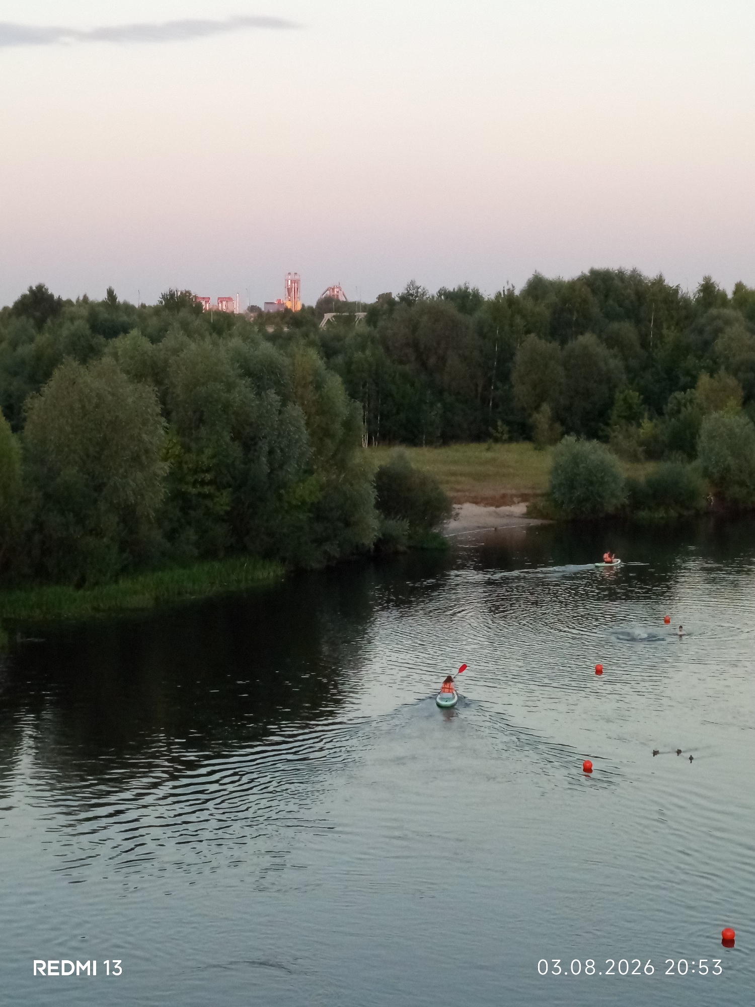 An aerial, wide shot captures a serene river scene in Barysaw, Belarus, during the late evening. The sky is a soft gradient of pale pink and blue, with a hint of industrial structures in the far distance. The river, a dark, rippling expanse, dominates the foreground and mid-ground. On the left bank, a dense, dark green forest stretches across the frame, with a thin strip of lighter green grass bordering the water. Further inland, beyond the trees, a sandy patch is visible.
Several individuals are engaged in water activities. In the mid-ground, a person in an orange life vest is kayaking, their paddle creating a wake. Nearby, two other people are in the water, seemingly swimming or splashing, with one visible head bobbing above the surface. A second kayaker, also wearing an orange life vest, is further upriver. Scattered across the water are several bright orange buoys, marking a course or designated area. Two ducks are also present, paddling calmly on the water. The timestamp "03.08.2026 20:53" and the text "REDMI 13" are visible in the bottom right and left corners respectively.