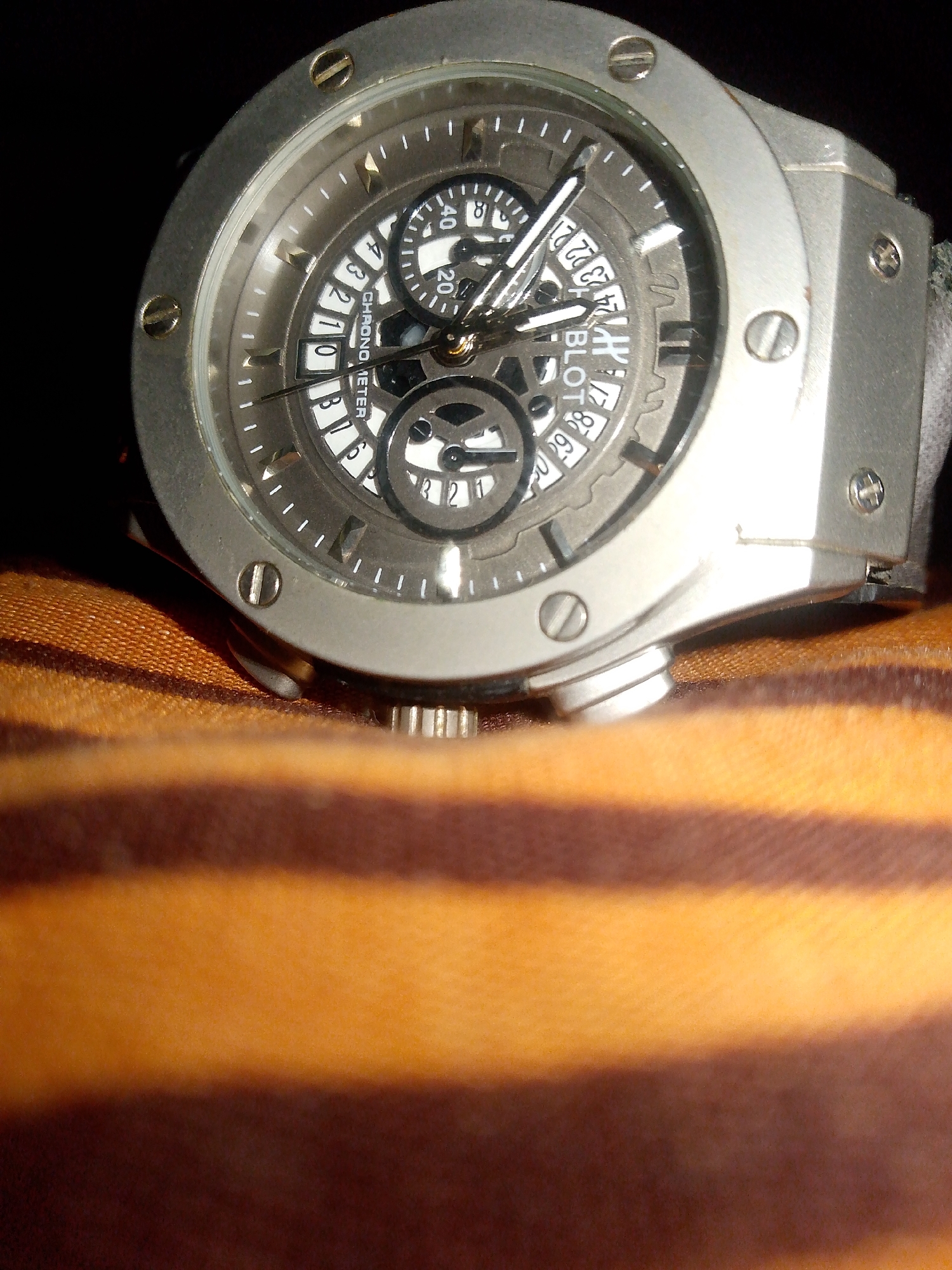 A close-up, slightly blurred, low-angle shot shows the face of a men's wristwatch. The watch has a silver-toned, hexagonal case with visible screws on the bezel. The watch face is dark gray with silver hour markers and white Arabic numerals. There are multiple subdials, one of which shows a date complication with a date window displaying "22". The word "CHRONOMETER" is visible below the date window. The watch hands are silver and pointed. The watch is lying on a surface with a striped pattern, appearing to be made of fabric, with alternating bands of orange and dark brown. The background is dark, suggesting low ambient light.