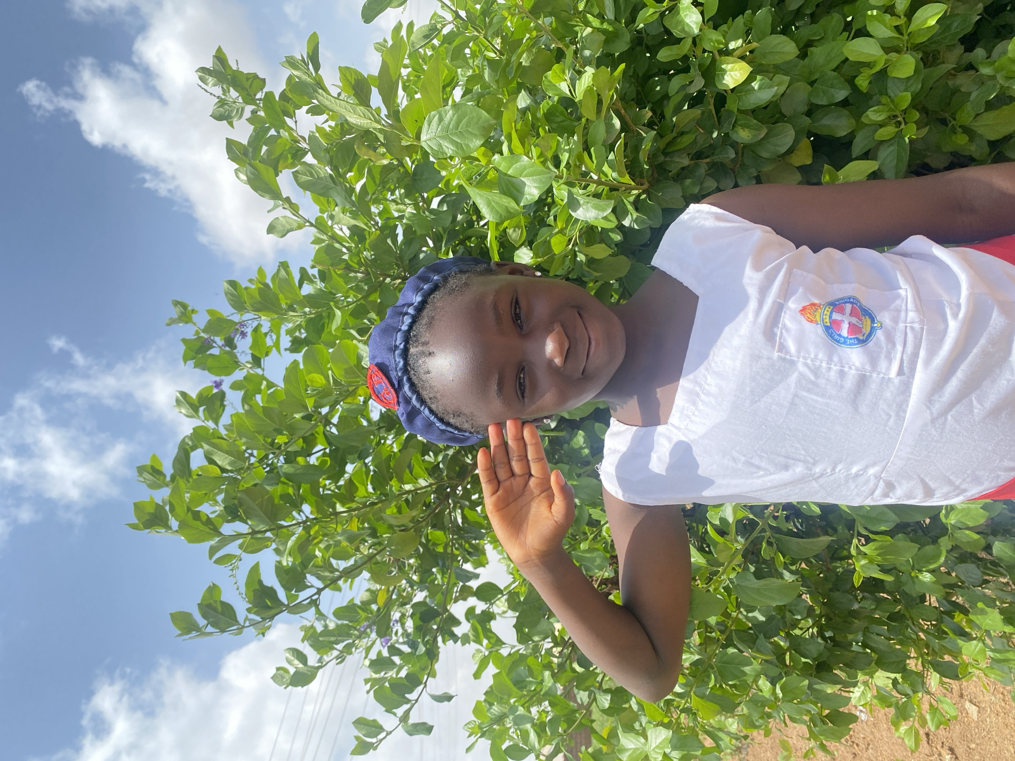 A young Black girl is standing outside in Turu, Nigeria. She is wearing a white shirt with a patch that says "The Girls Brigade" and a blue hat. She is smiling and waving her hand towards the camera. Behind her, there is a green leafy shrub and a blue sky with white clouds. The ground appears to be dirt.