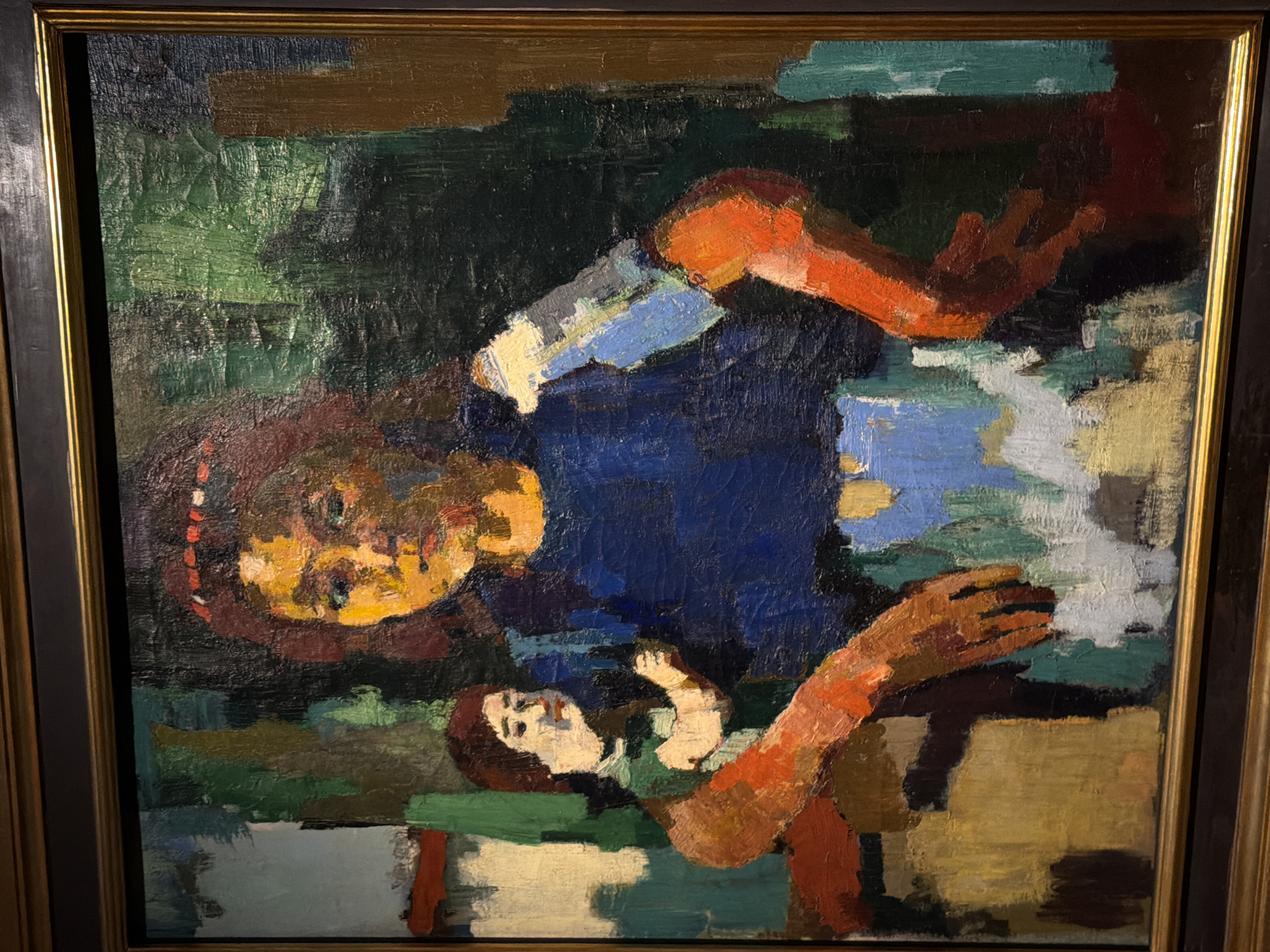 The image is a framed oil painting, displayed in an indoor setting. The painting depicts two figures, likely a mother and child, rendered in a semi-abstract, expressionistic style with bold brushstrokes and vibrant colors. The primary figure, presumably the mother, is positioned centrally, facing forward with her head slightly tilted. She has a dark red hairband with what appear to be small red and white beads, a dark blue garment, and prominent orange-red arms. Her face is painted in yellows and oranges, with dark eyes and a reddish mouth. Her left arm is bent and rests on a surface, while her right arm is also bent, with her hand visible. A smaller figure, the child, is positioned to the mother's left, seen in profile, wearing a green top and orange-red clothing. The background is a mixture of dark greens, blues, and browns, with some lighter abstract shapes suggesting a backdrop. The painting is housed in a dark wooden frame with a thinner gold inner frame. The lighting suggests overhead illumination, casting soft shadows. The location is a museum or gallery, evidenced by the framing, lighting, and presentation of the artwork. It is highly unlikely this painting is located in the city of Seoul, South Korea, based on the visible context.