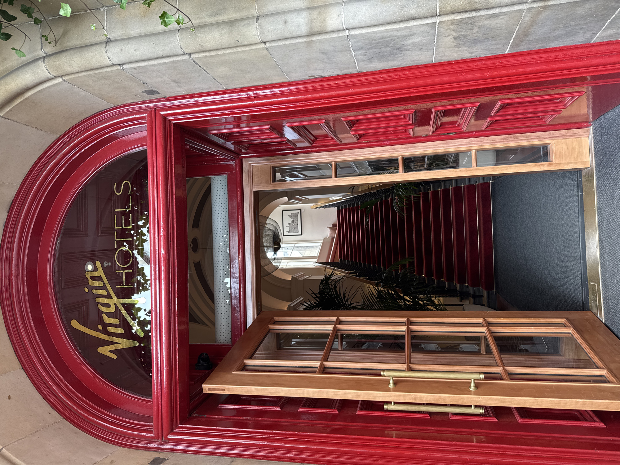 A full exterior shot captures the grand entrance of Virgin Hotels Edinburgh. The facade features a large, arched red doorway with ornate trim. Above the doorway, a semicircular glass pane displays the hotel's name in gold lettering: "Virgin HOTELS". The main door is ajar, revealing a glimpse of the interior. To the left, a double wooden door with glass panes and brass handles stands open, offering a view of a carpeted staircase. The staircase, lined with red carpet and brass accents, ascends into the building. A large potted plant is visible to the left of the stairs. The surrounding architecture is of stone masonry.