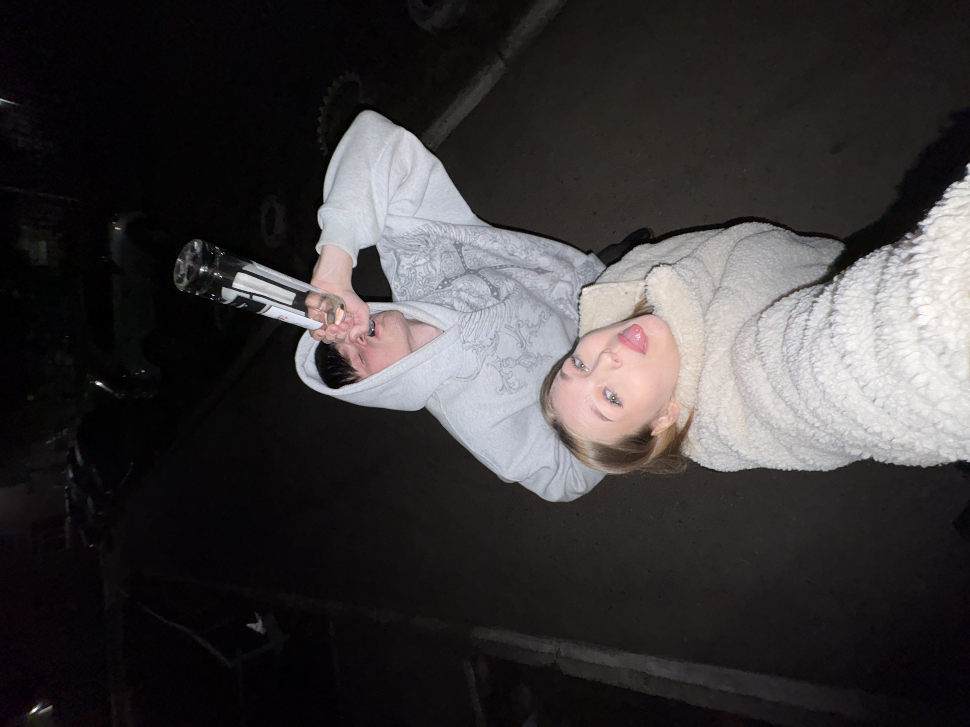 A photo taken outdoors in a dimly lit environment shows two individuals, likely posing for a selfie. A fair-skinned young woman is wearing a cream-colored, plush, sherpa-type jacket. She is facing the camera with her tongue slightly sticking out. Next to her is a fair-skinned young man wearing a light grey hooded sweatshirt with a graphic design on the front. He is holding a clear glass bottle. The surroundings are dark, with hints of urban elements like asphalt and indistinct structures. It is not possible to confirm that this image was taken in the city of Chaikovsky, Russia.