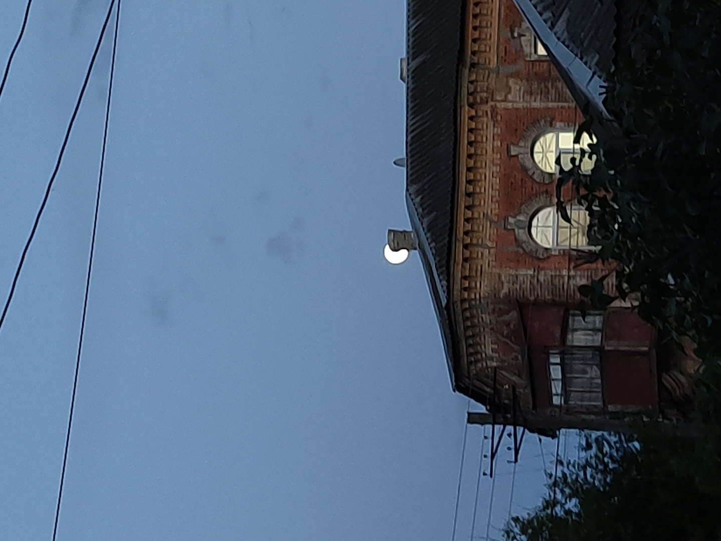 The image shows a building facade at dusk, with a streetlight illuminating the structure. The building is made of red brick, and has several windows, including two circular ones on the upper level. Power lines run along the left side of the frame, and trees partially obscure the right side of the building. The location is described as Unknown, Unknown.