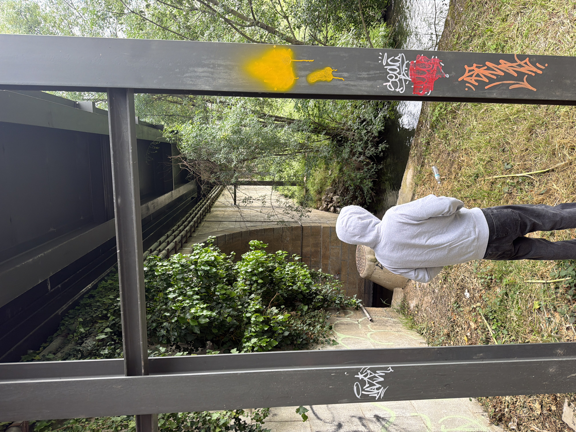 A man wearing a grey hoodie stands beside a storm drain, near a railing covered in graffiti. The railing is painted dark grey and has yellow, red, white, and orange graffiti on it. Beyond the railing, ivy grows on a stone structure. A stream runs beneath the stone structure. The image was taken in Bragança, Portugal.
