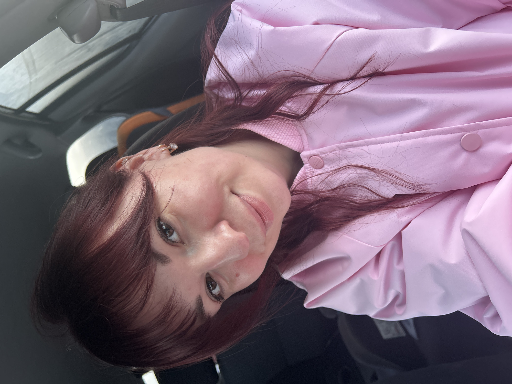 A woman with shoulder-length auburn hair is seated in a car, wearing a light pink jacket. She looks directly at the camera with a slight smile. The car interior is visible, including the roof and a glimpse of the passenger seat.