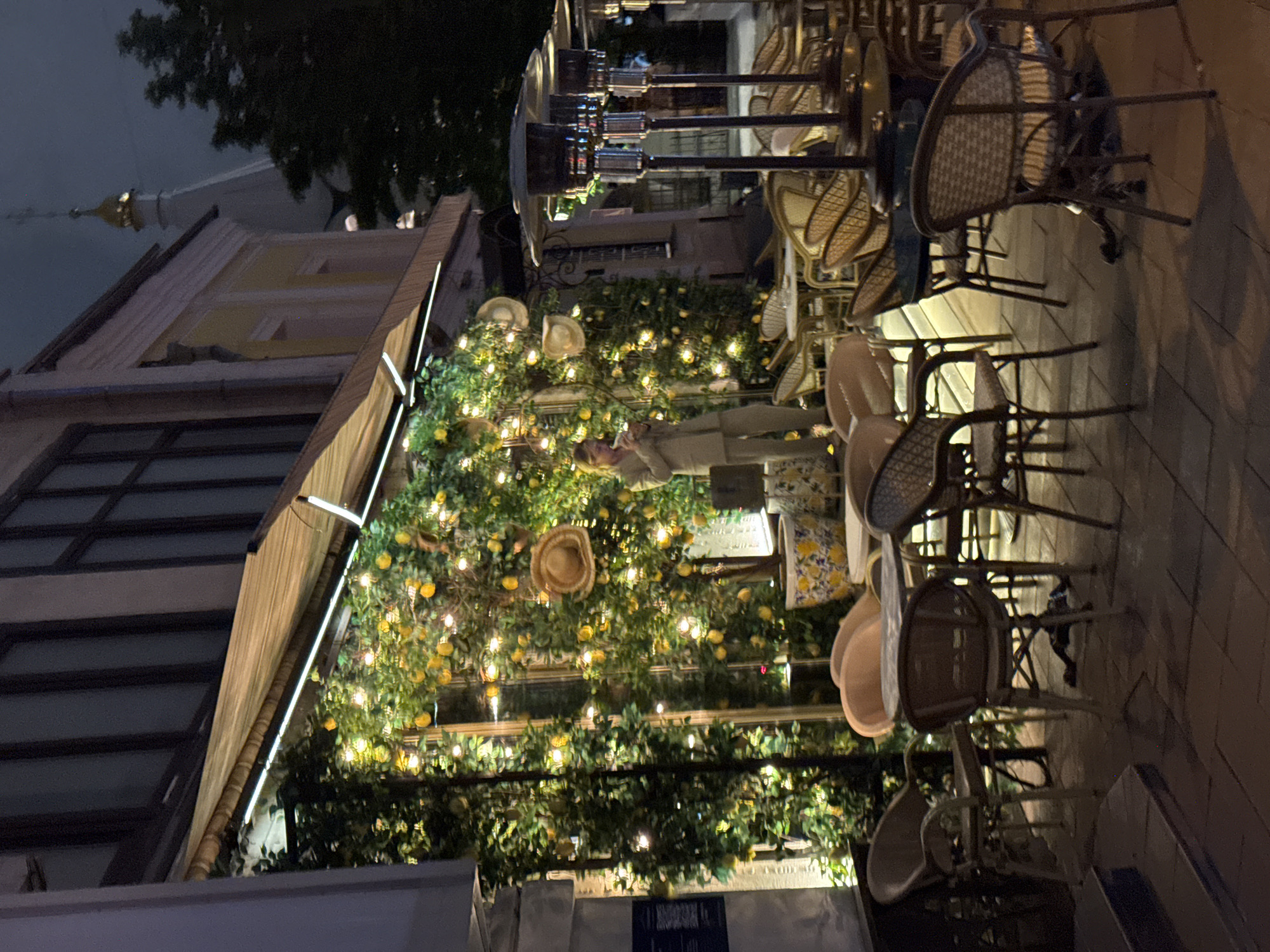 This outdoor scene shows an urban cafe in Moscow, Russia at night. The cafe is decorated with greenery, including lemon trees and string lights, under a striped awning. Straw hats are incorporated into the decor. A woman in a beige outfit is visible near the decorated wall. Adjacent to the cafe is a sidewalk with tables and chairs. In the background, the architecture of Moscow, including a church tower, is visible.