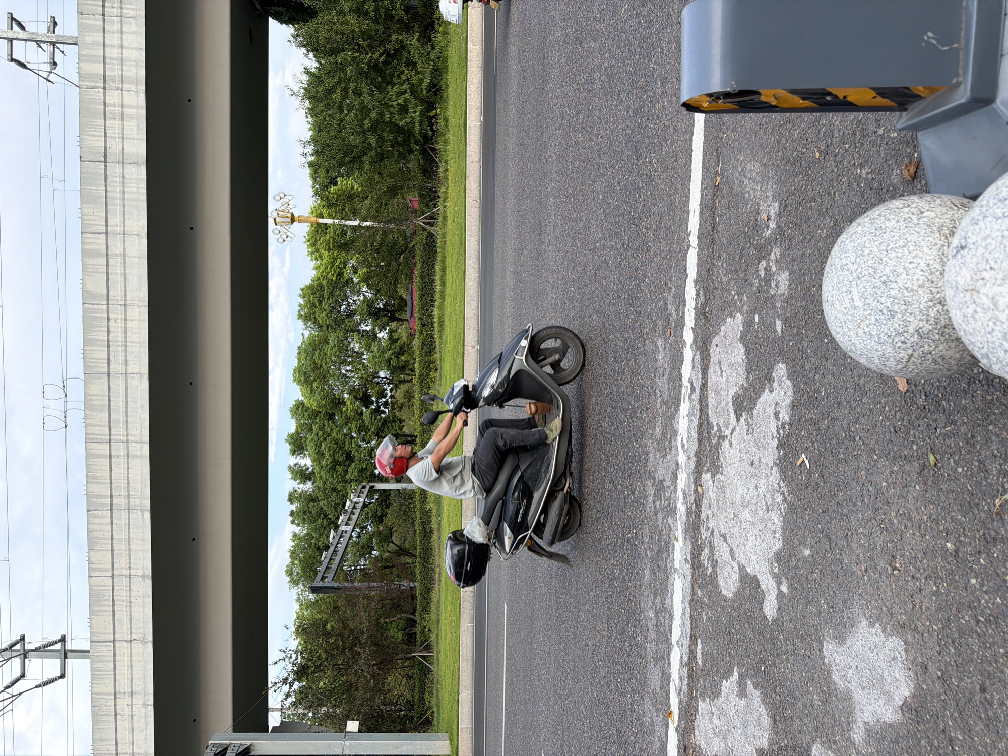 A man wearing a red helmet rides a black scooter on a paved road. The scooter has a black storage box on the back. The man is wearing a grey t-shirt and dark pants. The road has a white line marking the lane. In the background, there are trees and a large concrete overpass with power lines. The scene appears to be in an urban or suburban area.