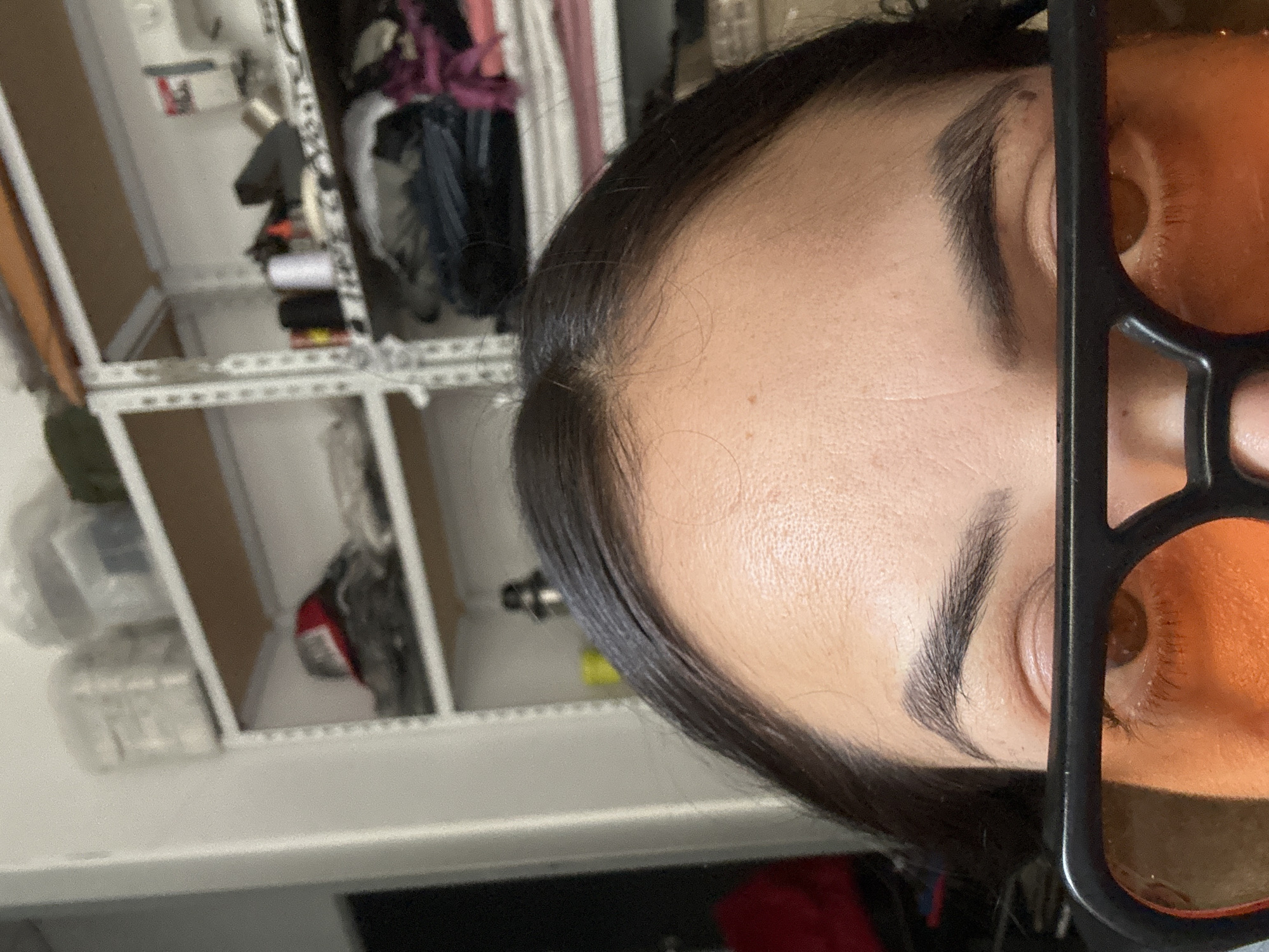 A close-up, rotated portrait shot focuses on a person's face, primarily the forehead, eyebrows, and one eye obscured by black-framed glasses with orange-tinted lenses. The eye appears to be brown. The hair is dark. The background features a closet or shelving unit with various items visible, including what appears to be clothing, and possibly storage containers. The location in Buenos Aires, Argentina is not deducible from the available image.