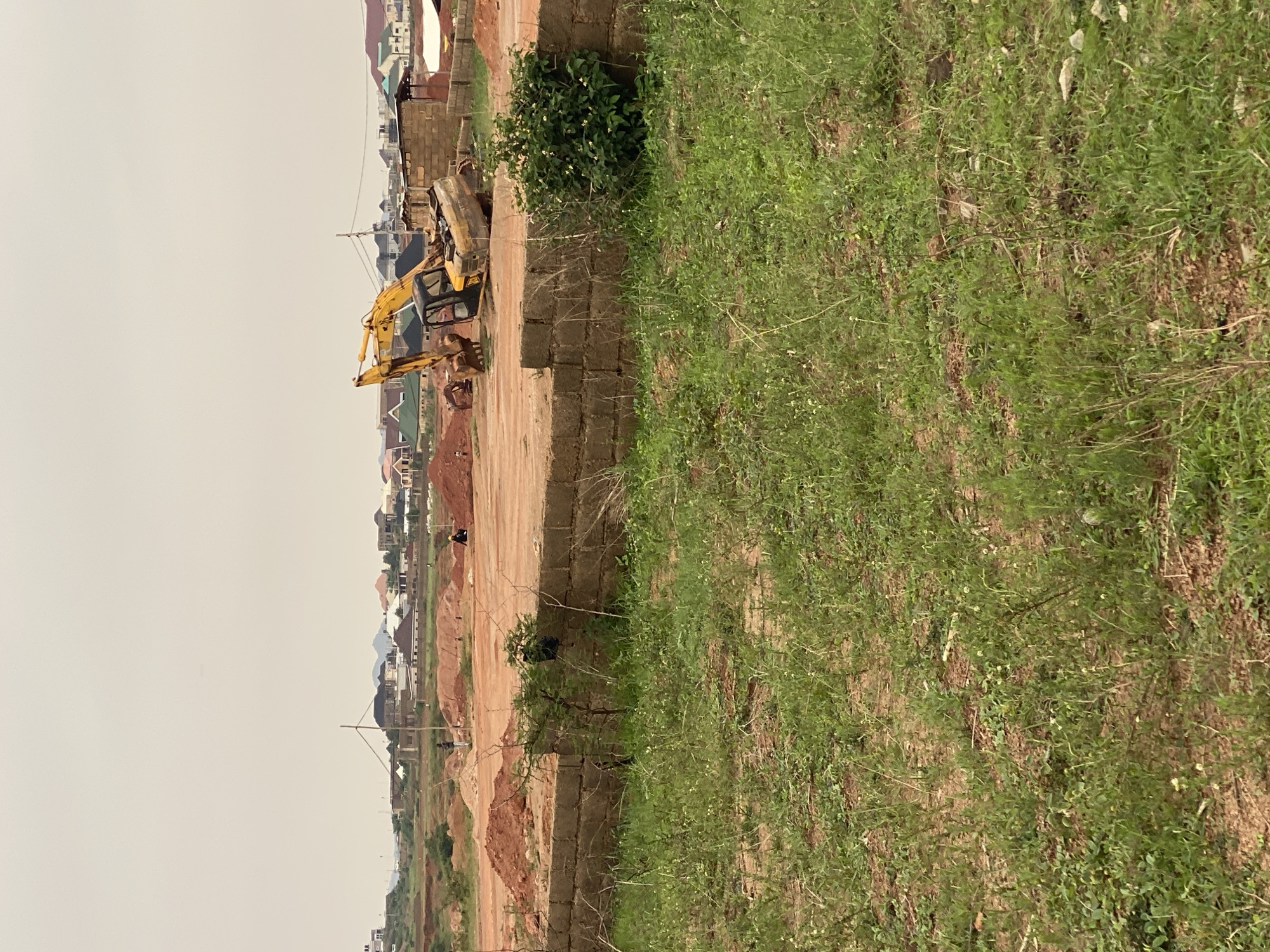 The image depicts a construction site in the city of Kwang, Nigeria, characterized by an overcast sky and a landscape dotted with unfinished buildings and mounds of red earth. In the mid-ground, a yellow excavator is parked. To the left of the excavator, two individuals are visible, one in dark clothing and the other in a yellow hard hat, standing near piles of dirt. Further in the background, a residential area with multiple houses, some appearing to be under construction and others completed, stretches across the horizon. The foreground is dominated by overgrown grass and a low brick wall.