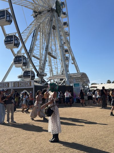 The image shows an outdoor scene in Indio, California, United States. The location appears to be a festival or event based on the presence of a large Ferris wheel, numerous people, and booths.
In the foreground, a person wearing a white dress, black boots, and a headscarf that obscures their face stands on dry, sandy ground. They have a black bag slung over their shoulder. Their shadow stretches out in front of them.
Behind them is a crowd of people dressed in casual clothing. Some individuals are using their phones, possibly taking pictures or videos. Behind the crowd is a booth or structure with "ID CHECK" printed on the side.
The background is dominated by a tall, white Ferris wheel against a clear blue sky. Other structures and tents are visible in the far distance. The overall scene suggests a festive and crowded environment under bright sunlight.